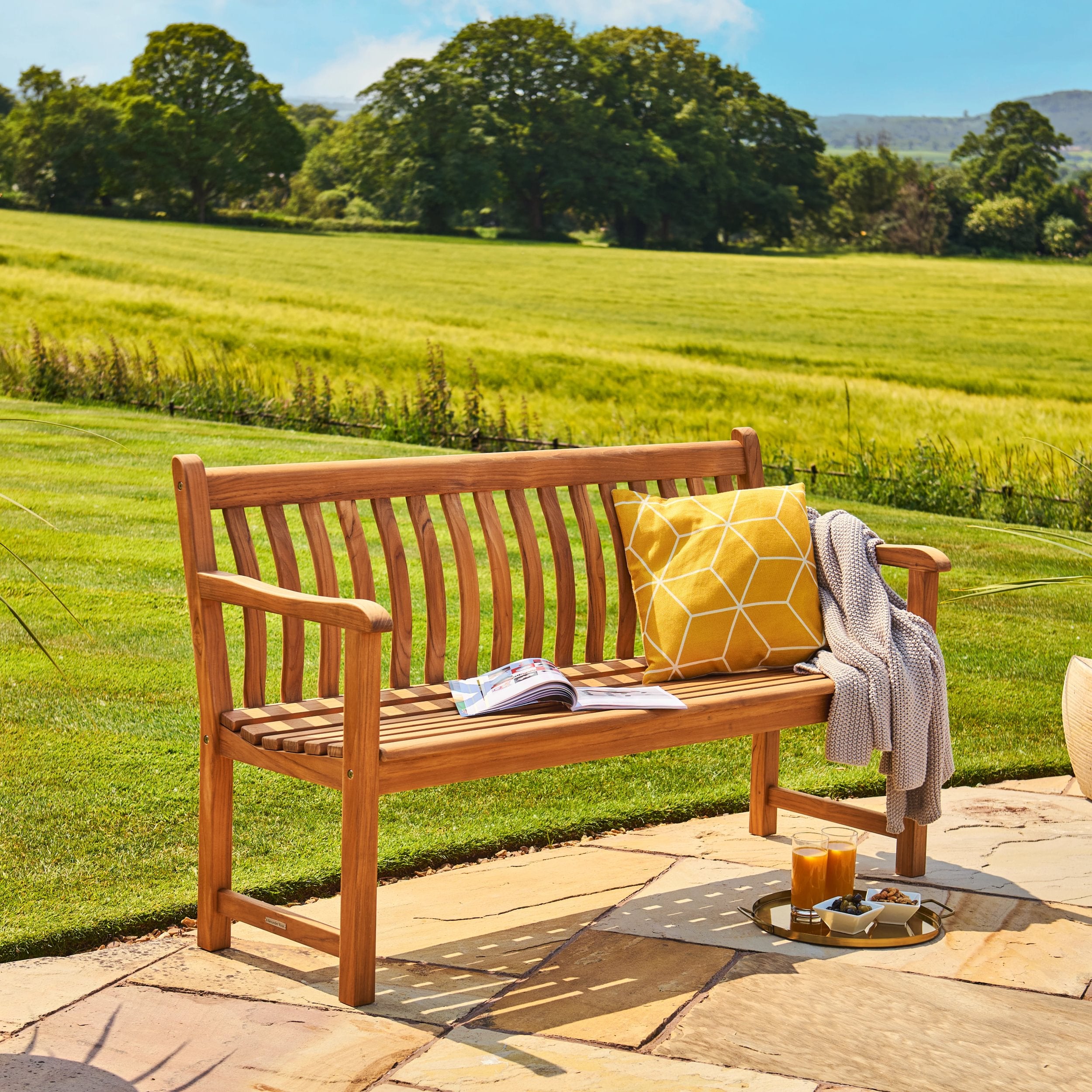 Wooden bench with a yellow cushion and a tray of items on a patio with a scenic background