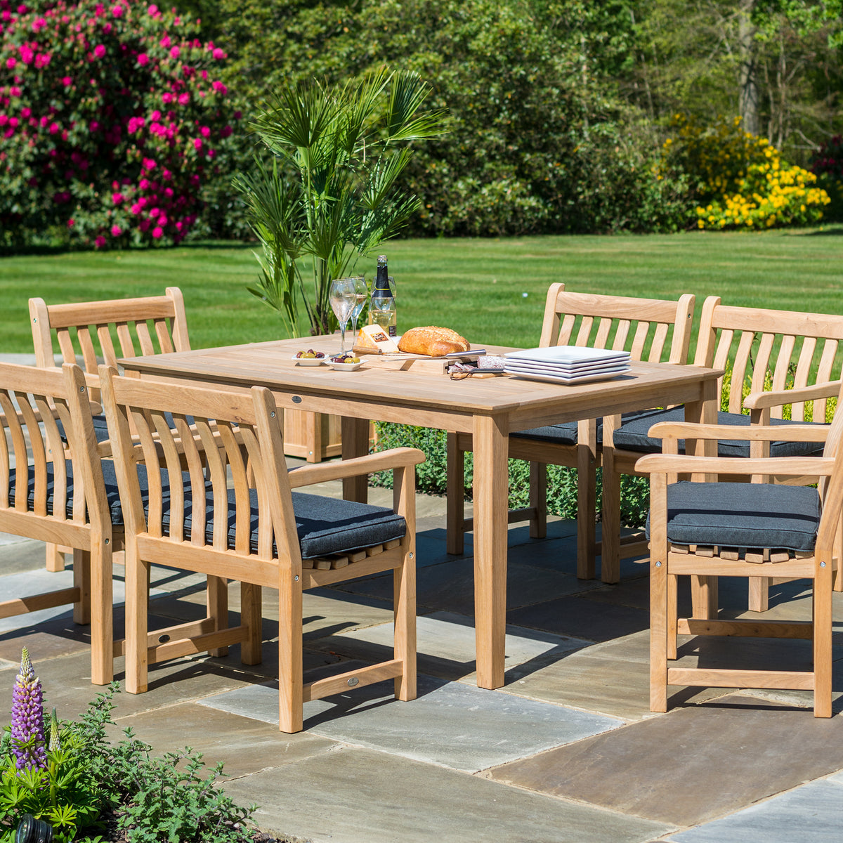 A 6-piece outdoor dining set consisting of a rectangular wooden table and six matching slatted armchairs with dark gray cushions, set on a stone patio in a lush green garden.