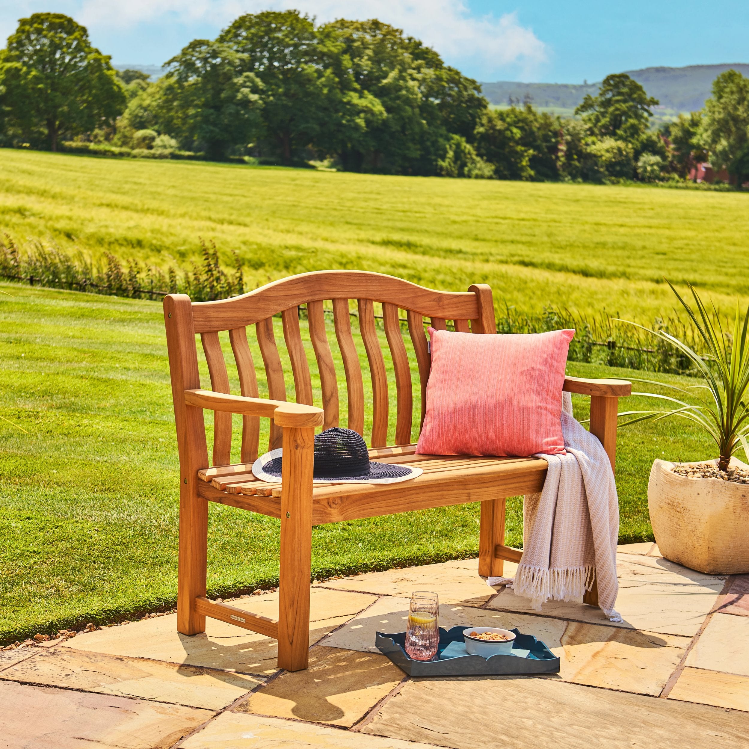 Wooden bench with a pink cushion and a hat on a stone patio with a scenic background of green fields and trees.