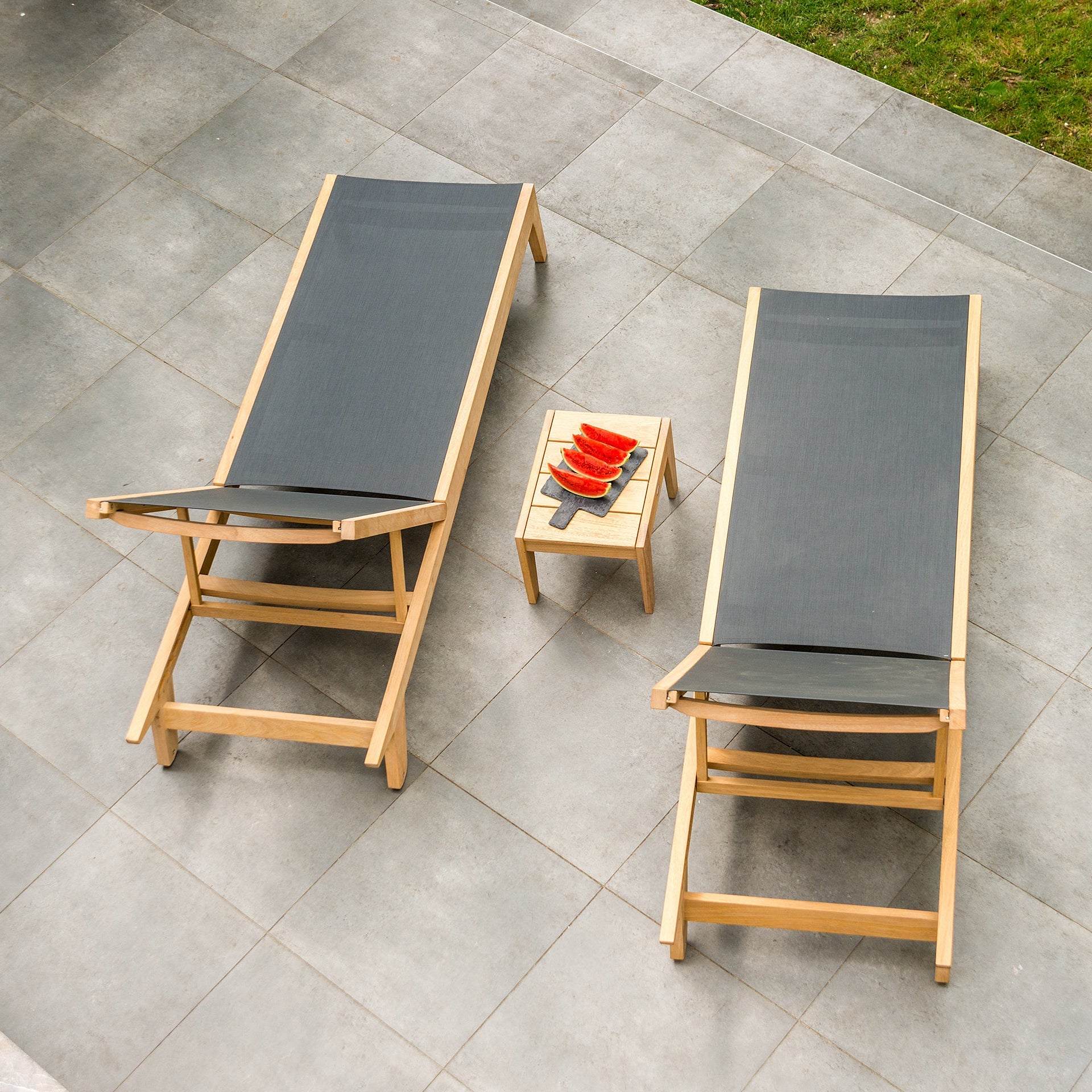 two slingback sun loungers with dark fabrics and wood frames on a patio with a small side table with melon
