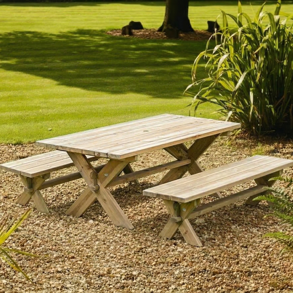 Wooden picnic table with benches on a gravel area with grass and plants in the background