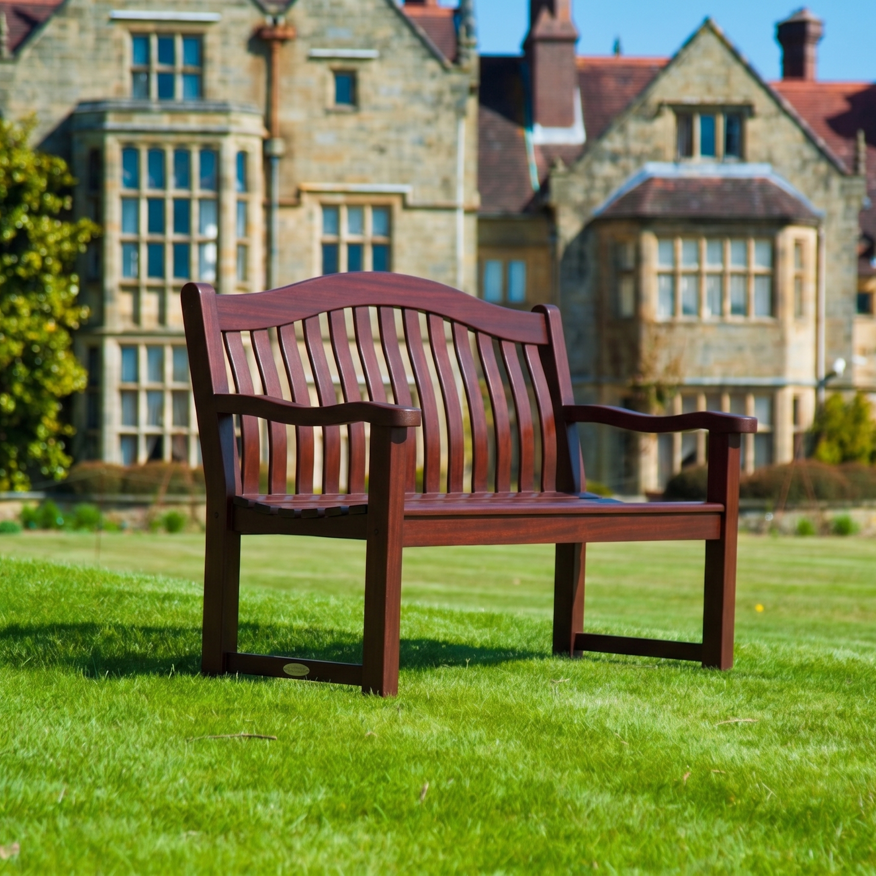 A mahogany wooden park bench with a slatted back and curved armrests, situated on a green lawn with a large stone building in the background.