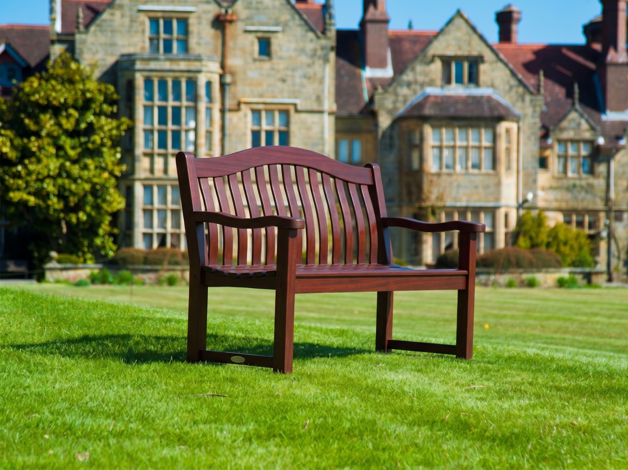 A mahogany wooden park bench with a slatted back and curved armrests, situated on a green lawn with a large stone building in the background.