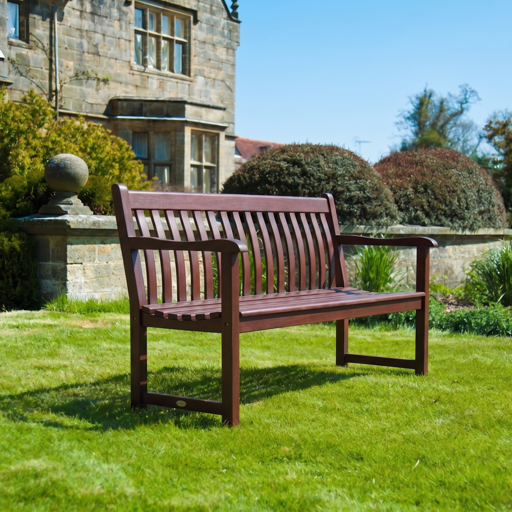 A mahogany wooden garden bench with slatted back and seat, placed on a green lawn in front of a stone building with shrubs and blue sky.