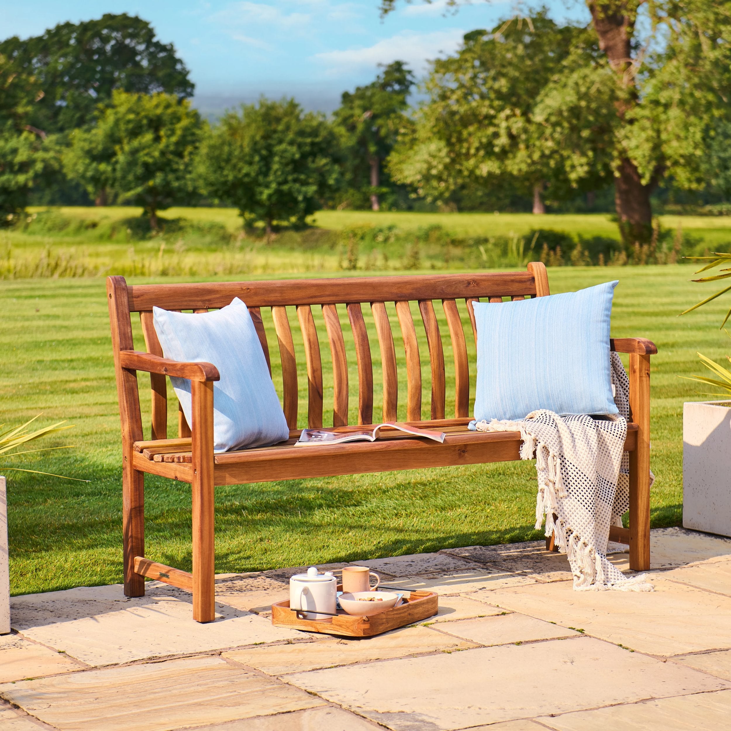 Wooden bench with blue pillows and a tray on a patio with a garden view