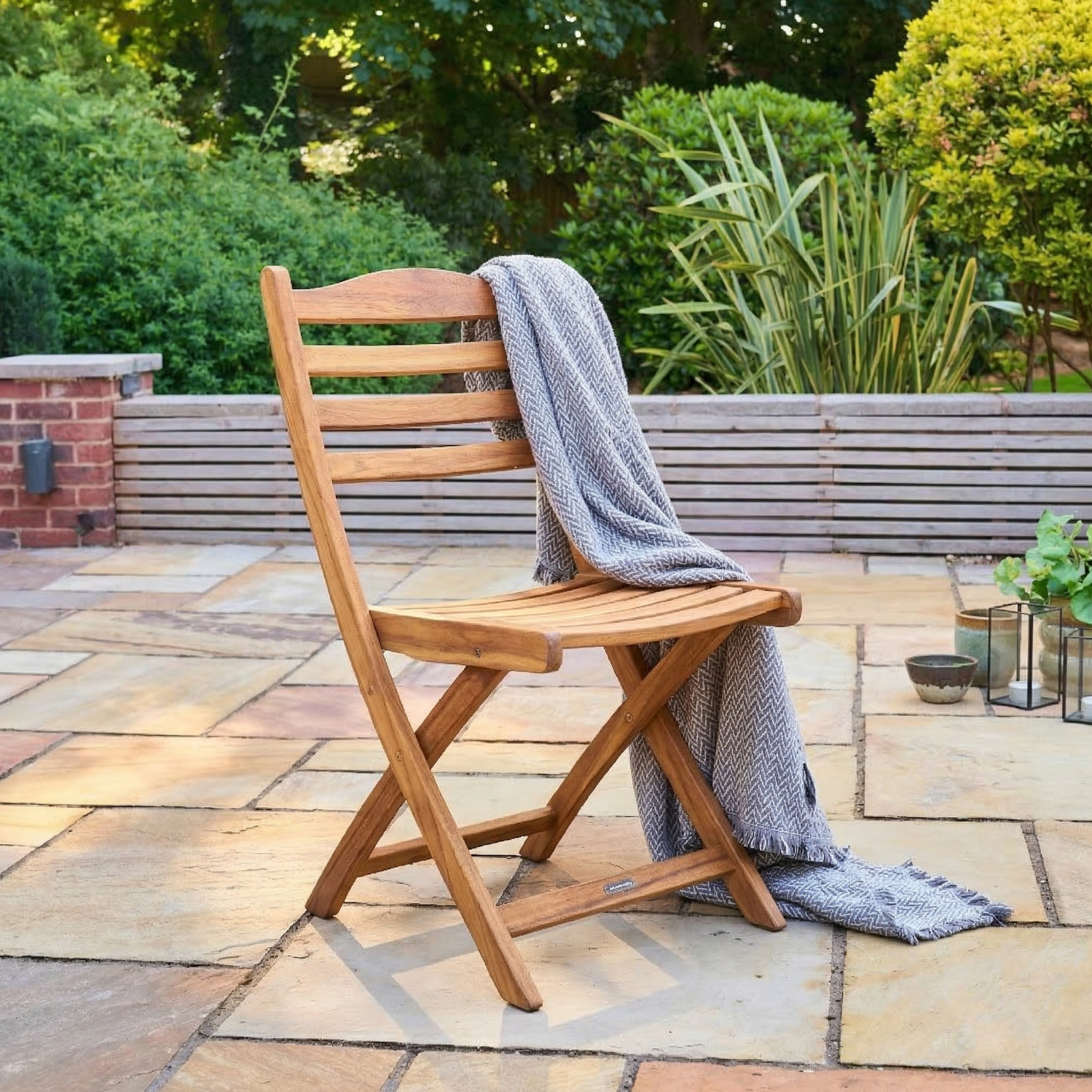 Wooden folding chair with a grey blanket on a stone patio with greenery in the background