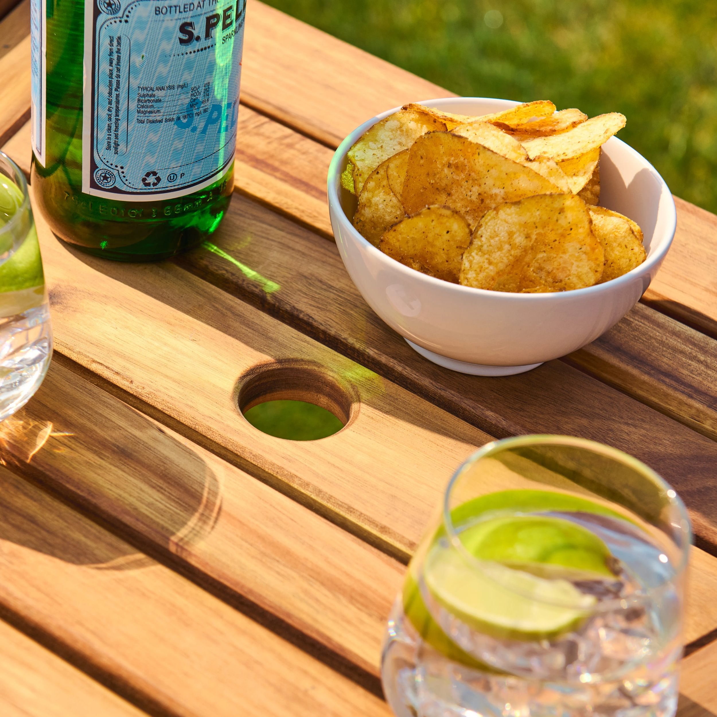 Bowl of crisps with a bottle and glasses on a wooden table surface