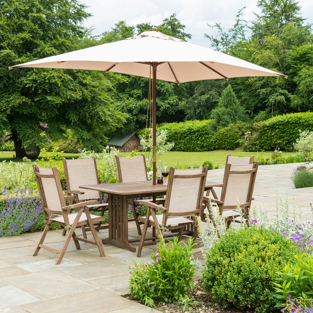 Garden setting with wooden dining table and sling recliner chairs under a large parasol, surrounded by greenery.