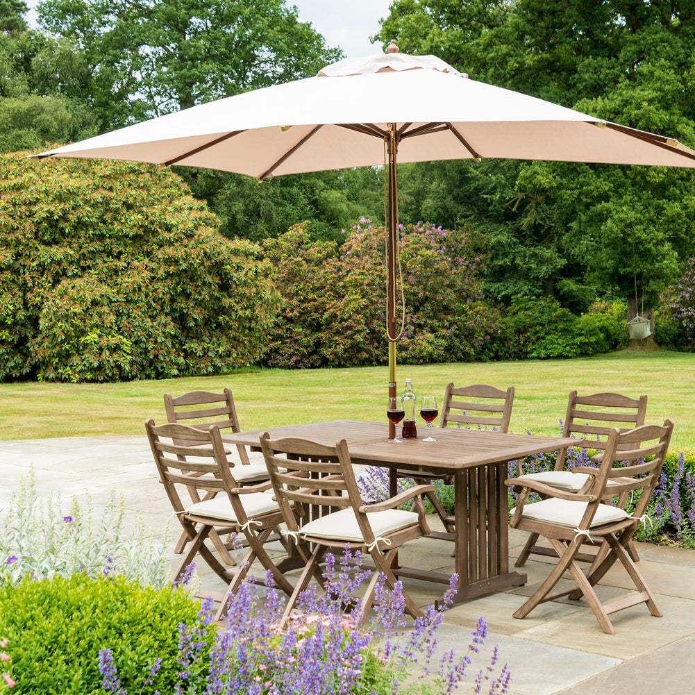 Garden setting with wooden dining table and chairs under a large beige umbrella, surrounded by greenery and flowers.
