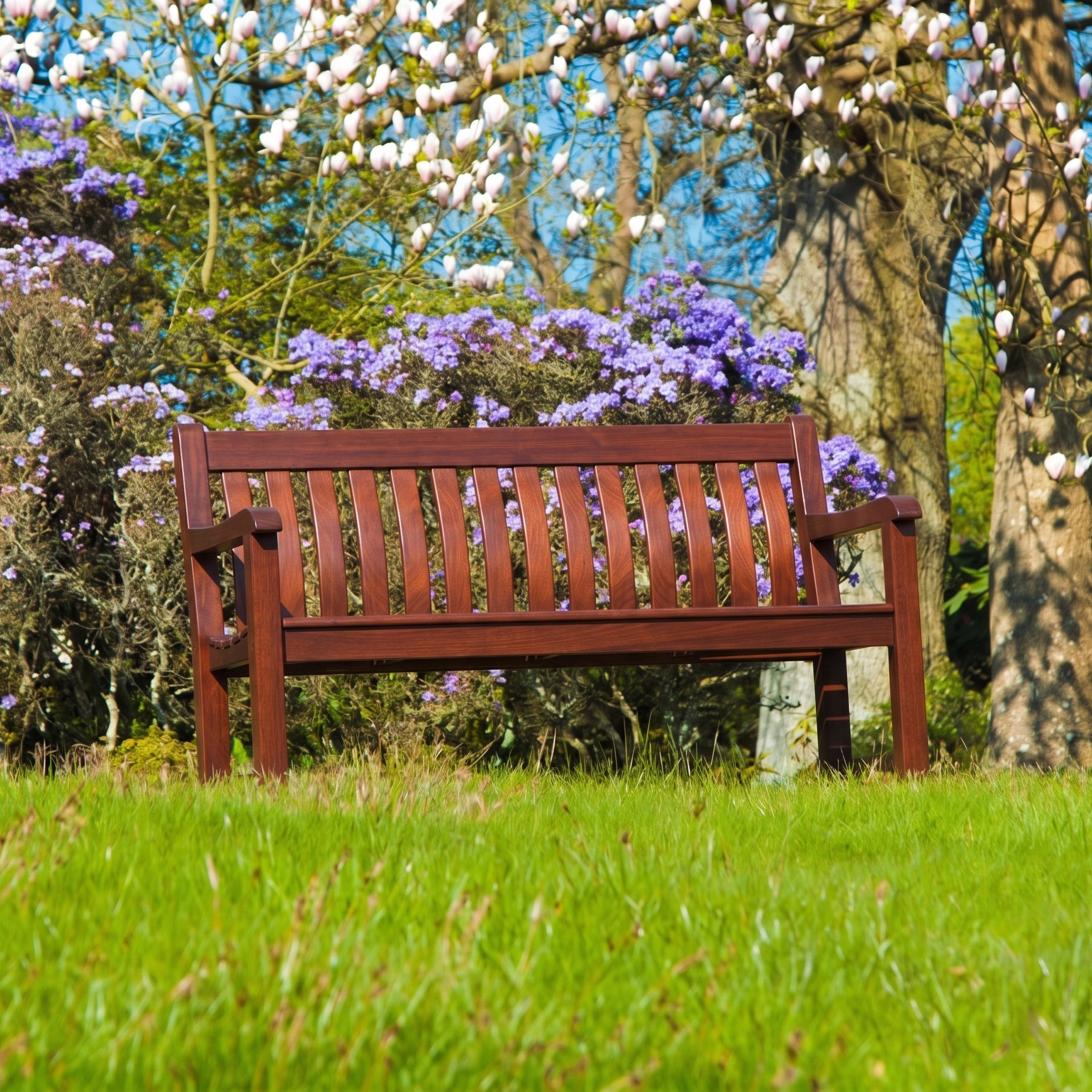A mahogany wooden bench situated in a grassy park area with blooming trees and purple flowers in the background.