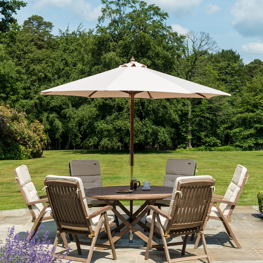 Outdoor patio set with table, 6 reclining chairs, and parasol on a stone patio with greenery in the background.