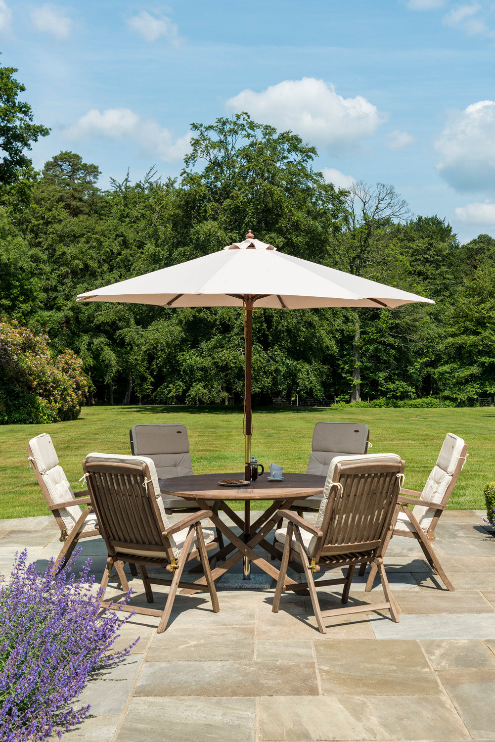 Outdoor patio set with table, 6 reclining chairs, and parasol on a stone patio with greenery in the background.