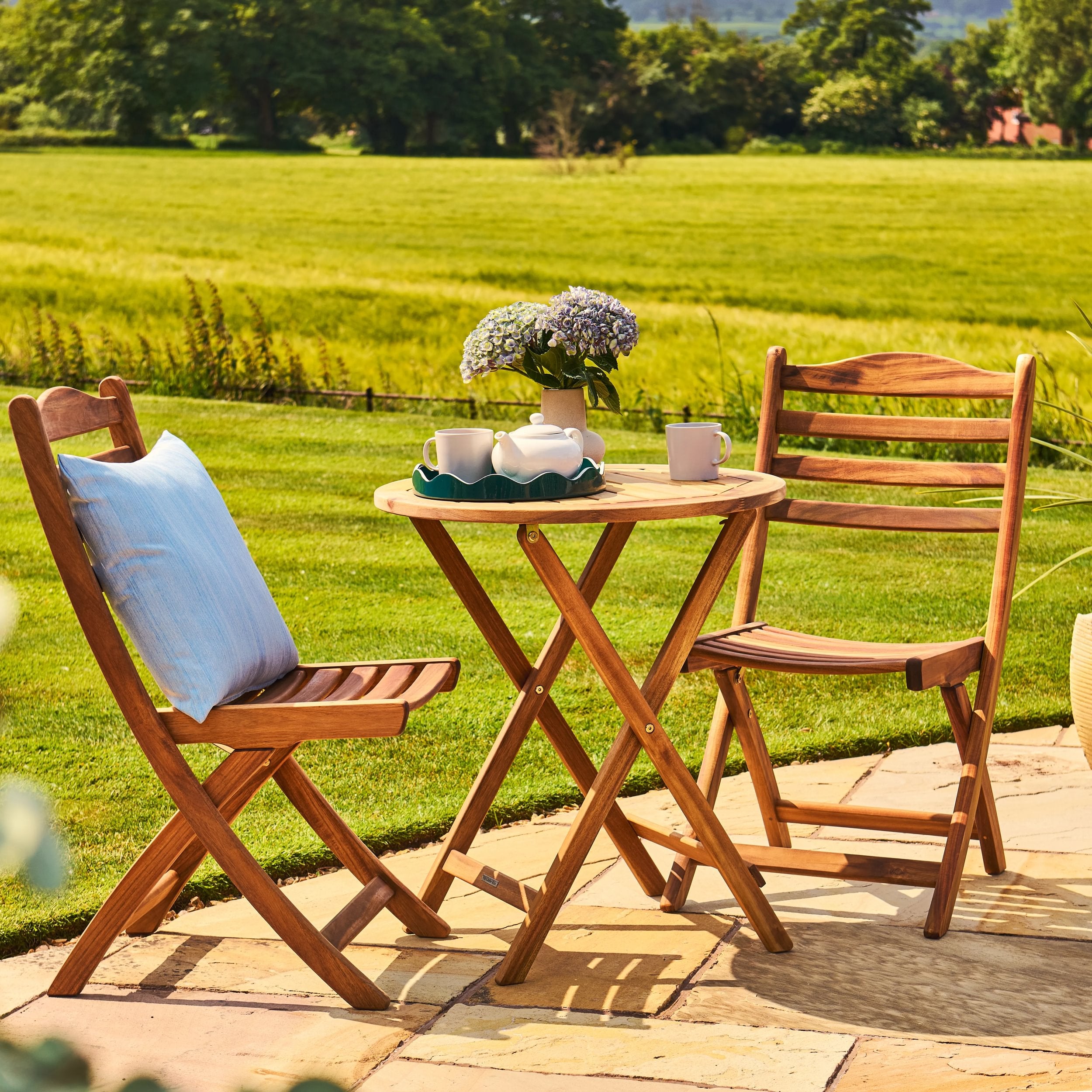 Wooden outdoor furniture set with a small table and chairs on a patio, surrounded by a green field.