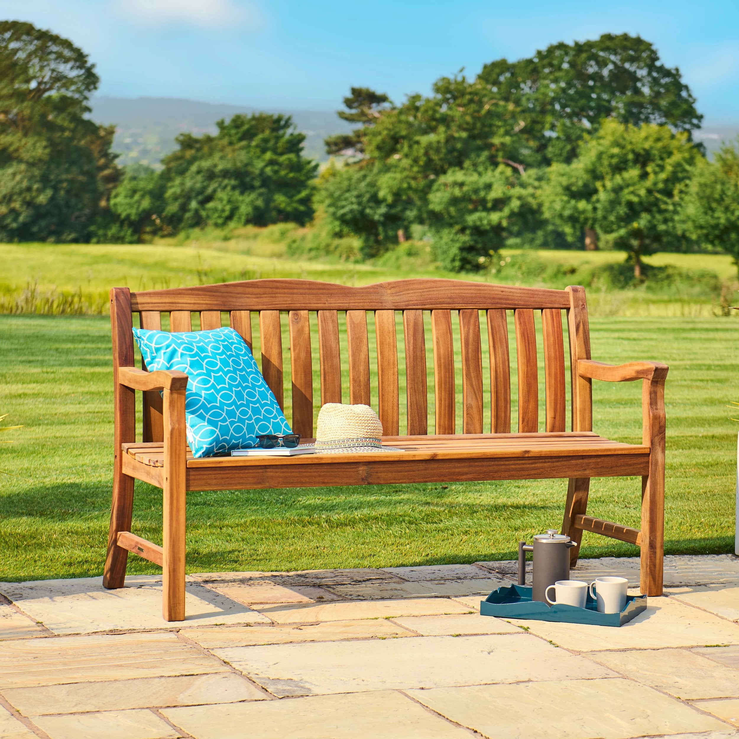 Wooden bench with a blue cushion and a tray of items on a patio with a green landscape in the background