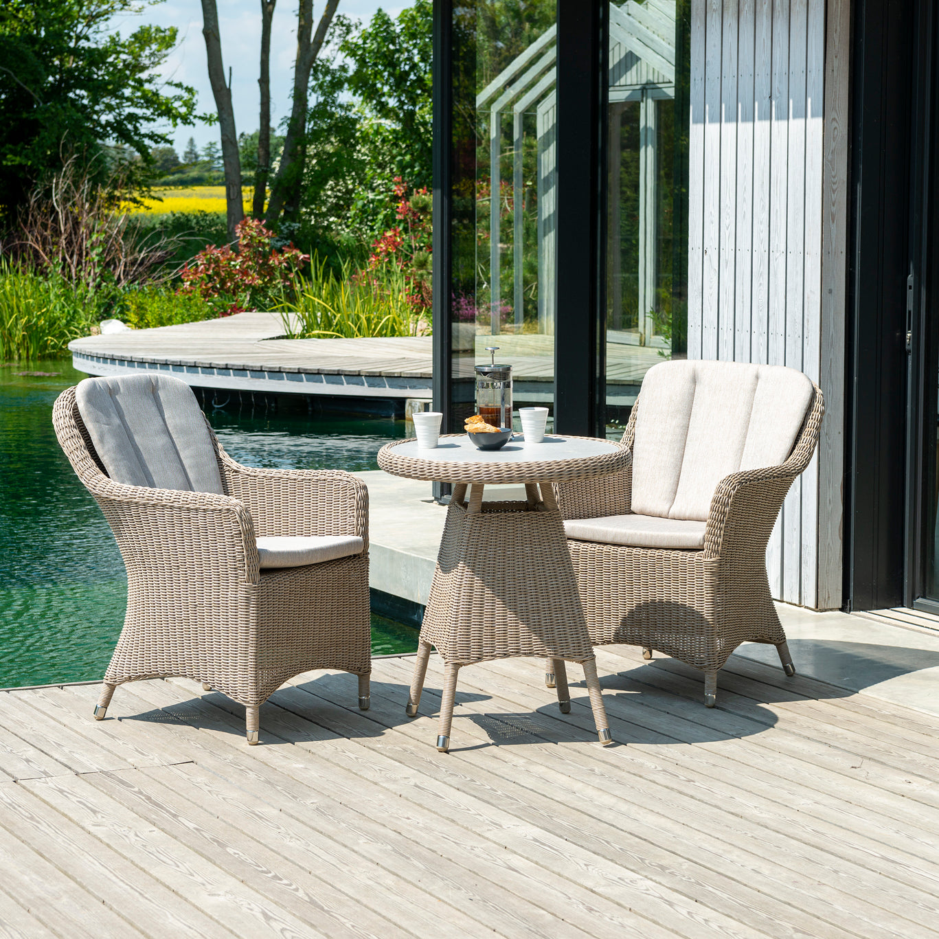 A three-piece outdoor patio furniture set featuring two beige wicker armchairs with light gray cushions and a matching round wicker side table, set on a wooden deck next to a swimming pool.