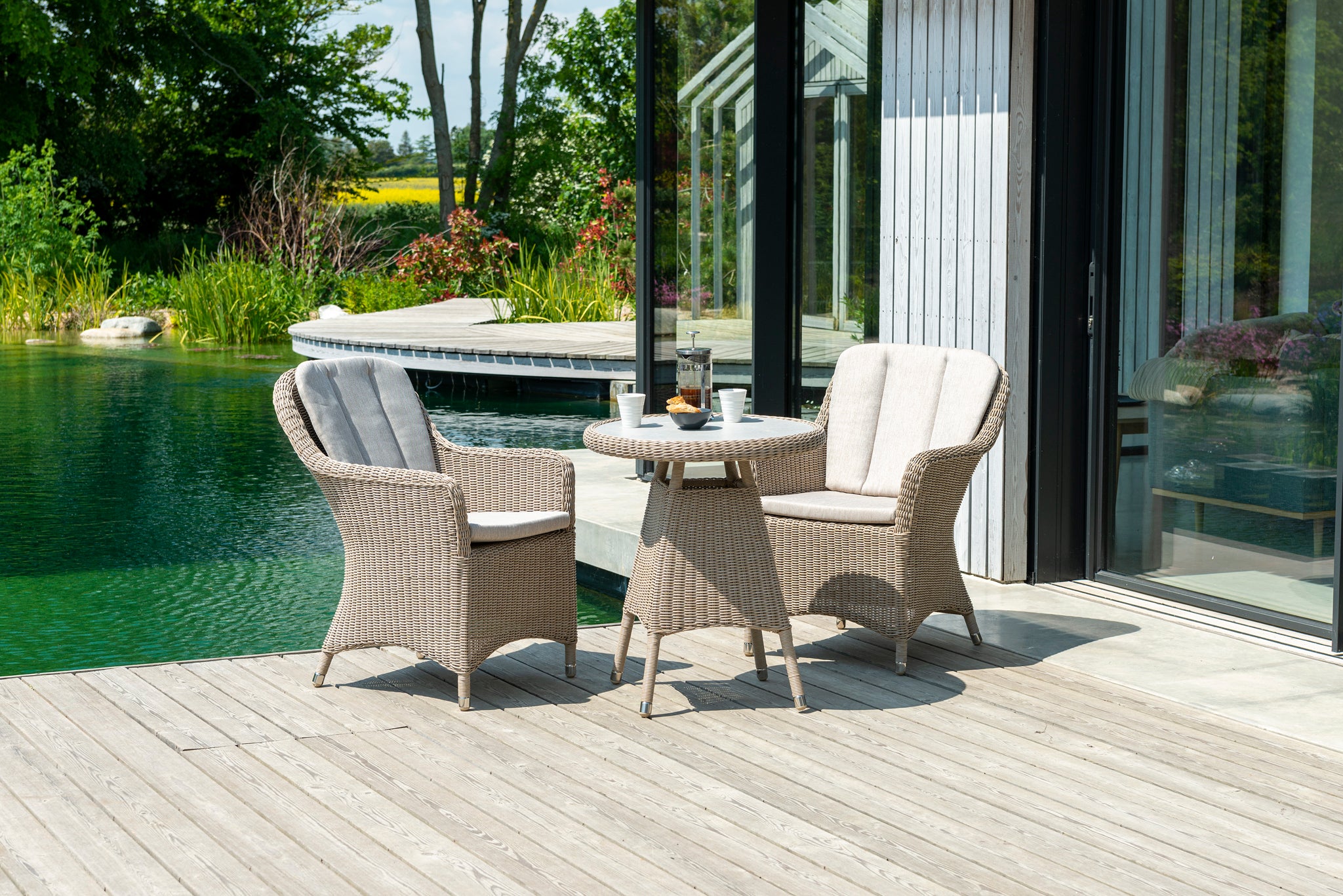 A three-piece outdoor patio furniture set featuring two beige wicker armchairs with light gray cushions and a matching round wicker side table, set on a wooden deck next to a swimming pool.