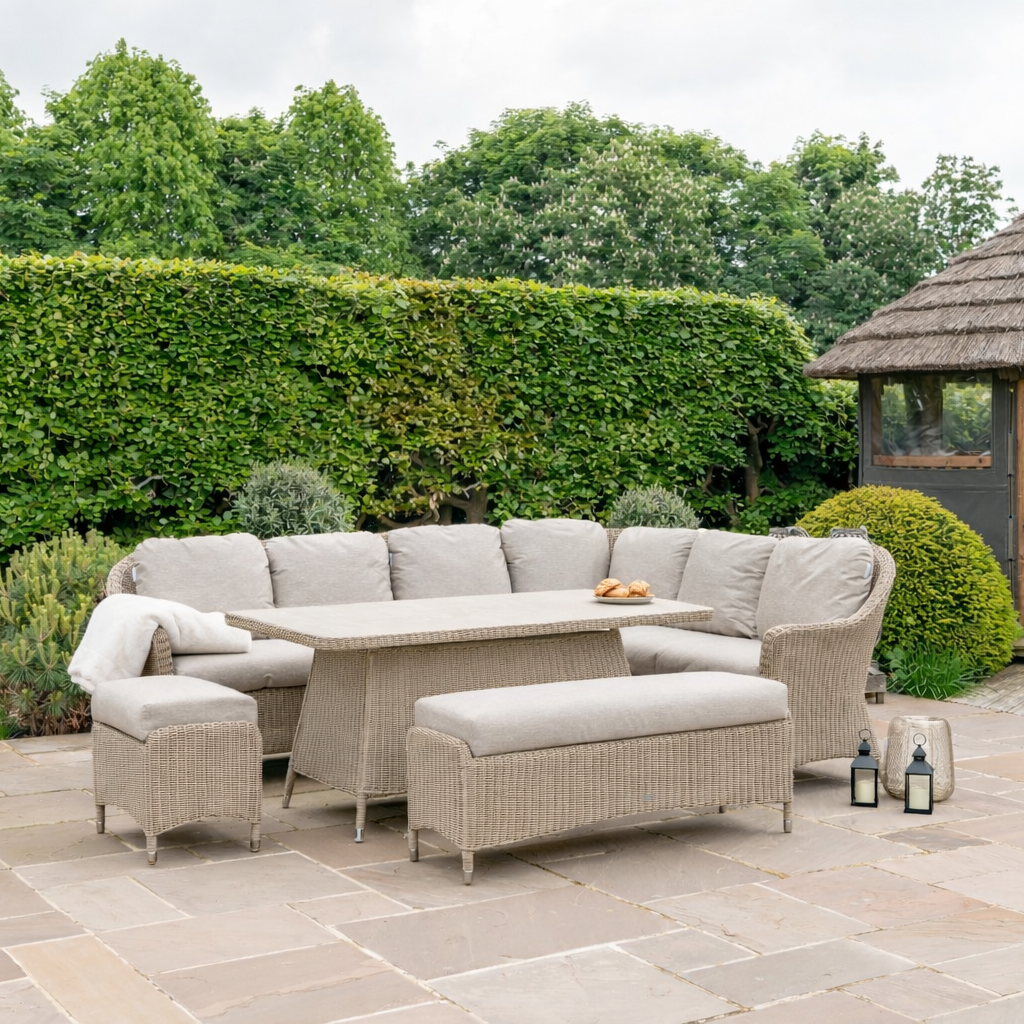 Outdoor patio furniture set with a curved sectional sofa, matching armchair, ottoman, and a rectangular wicker coffee table, arranged on a stone patio in a landscaped garden with a hedge and a small gazebo.