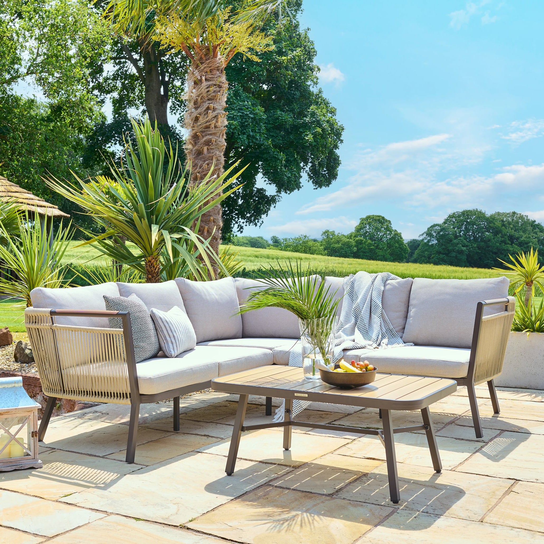Outdoor patio set with sectional sofa, chairs, and coffee table on a stone patio with greenery in the background.