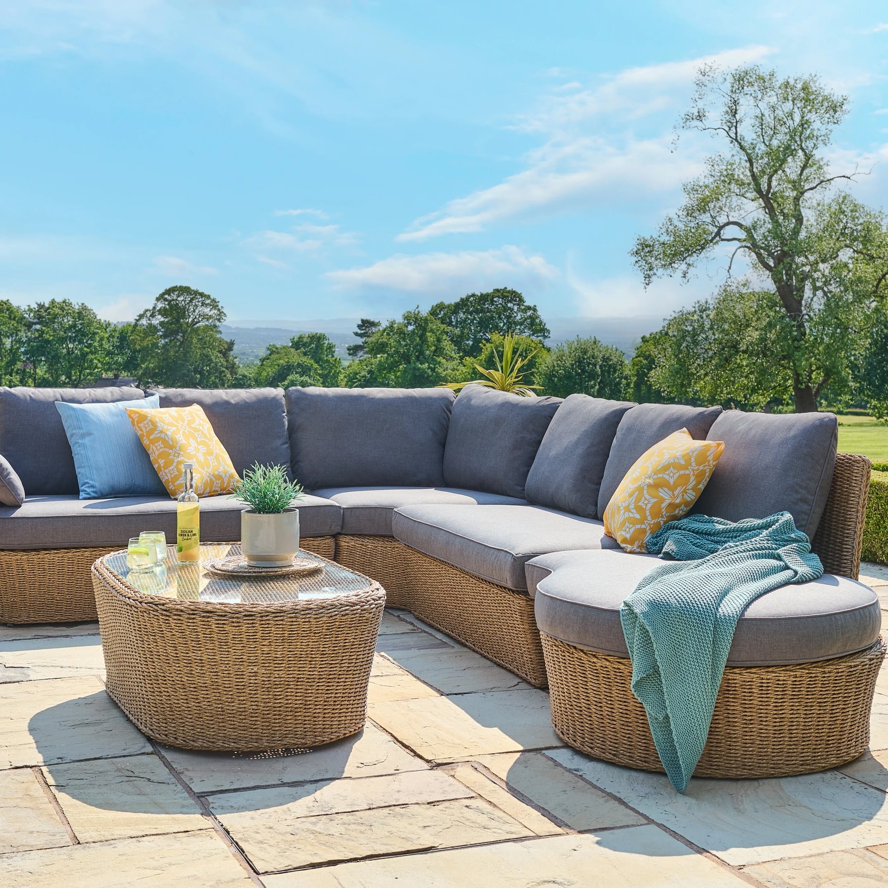 Outdoor patio furniture set with a sectional sofa and round table on a stone patio with a scenic background.