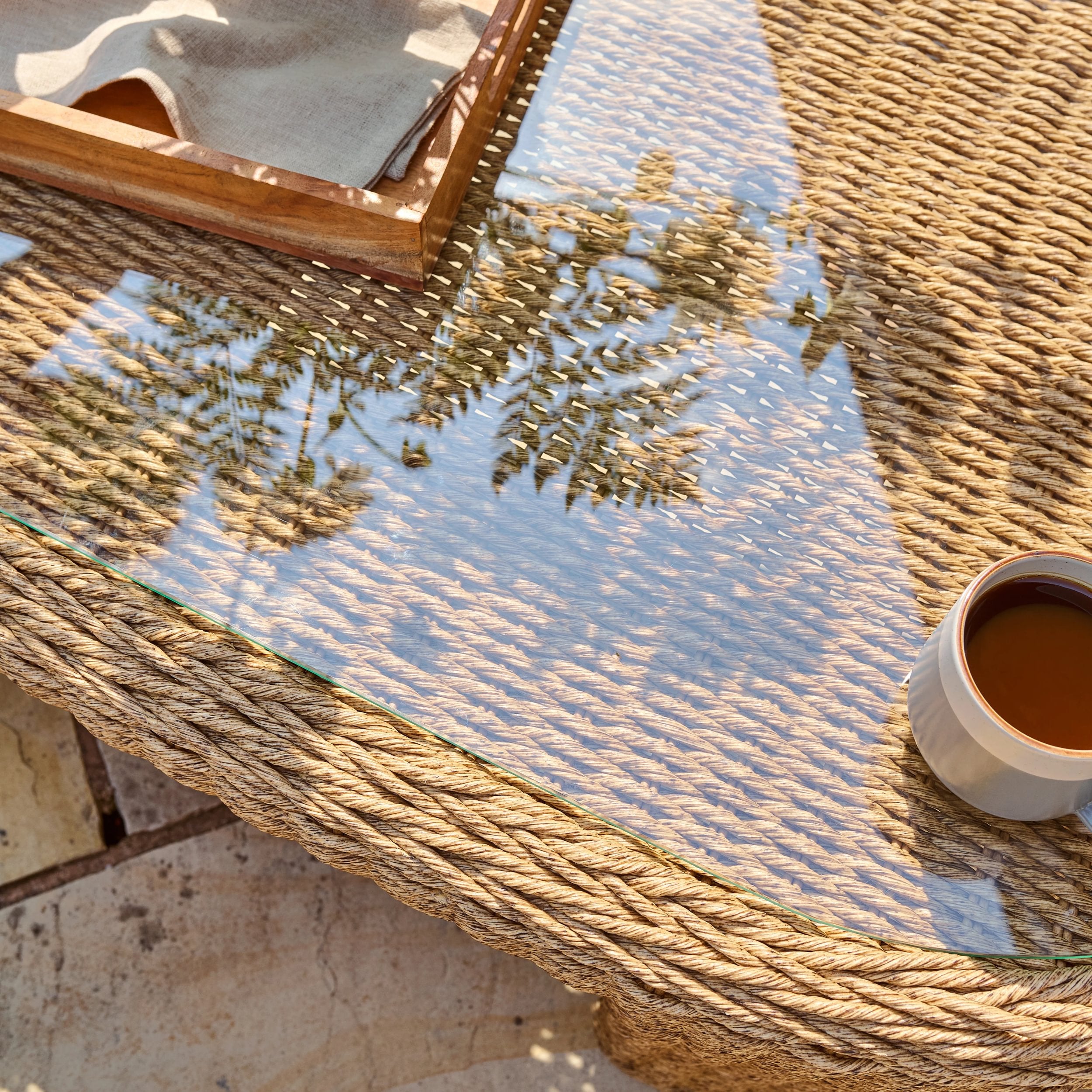 woven garden table with glass top and a wooden tray with a cup of coffee on a stone patio