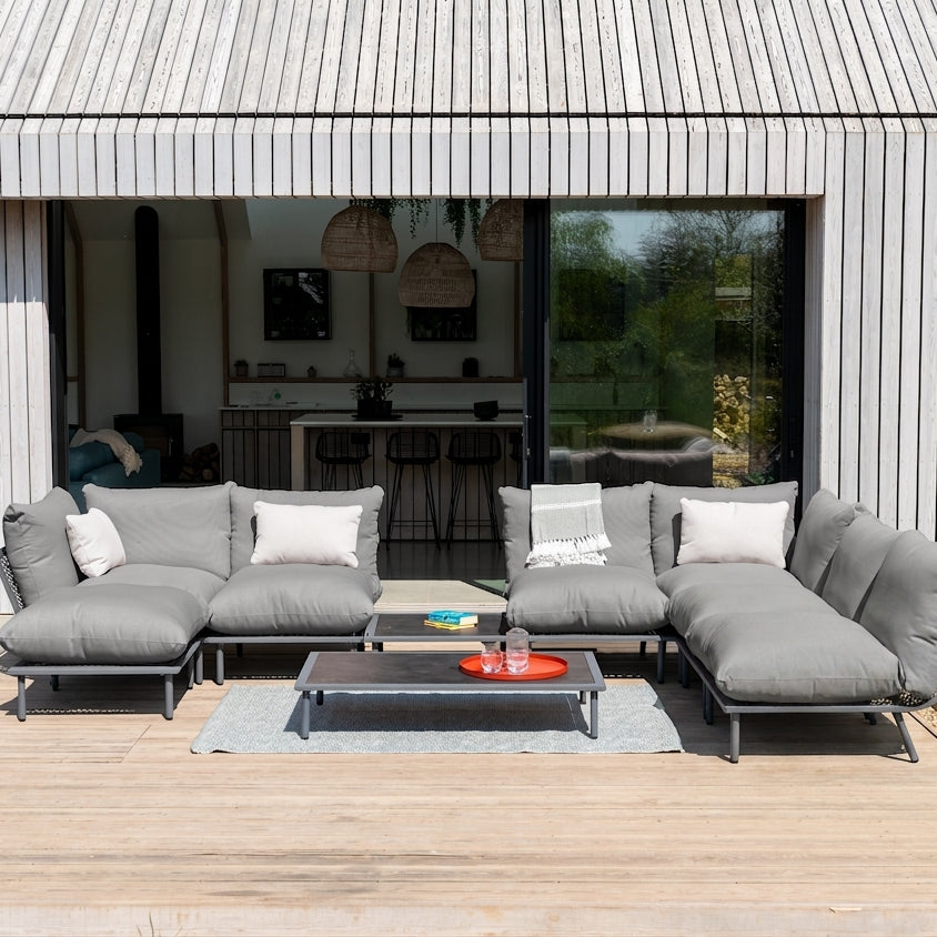 A modern outdoor sectional sofa set in gray fabric with white accent pillows, arranged on a wooden deck in front of a contemporary house with vertical siding and large glass windows.
