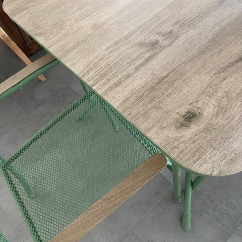 A close-up of a dining table with a light gray wood-grain top and mint green metal legs, alongside matching mint green mesh chairs.