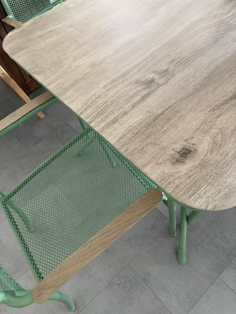 A close-up of a dining table with a light gray wood-grain top and mint green metal legs, alongside matching mint green mesh chairs.