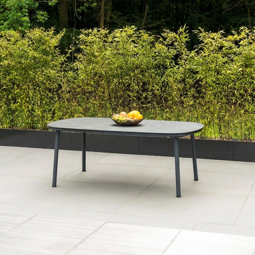 Outdoor patio table with pebble grey top. Rectangular with curved corners, on a tiled patio area with greenery in the background and a bowl of fruit in the centre