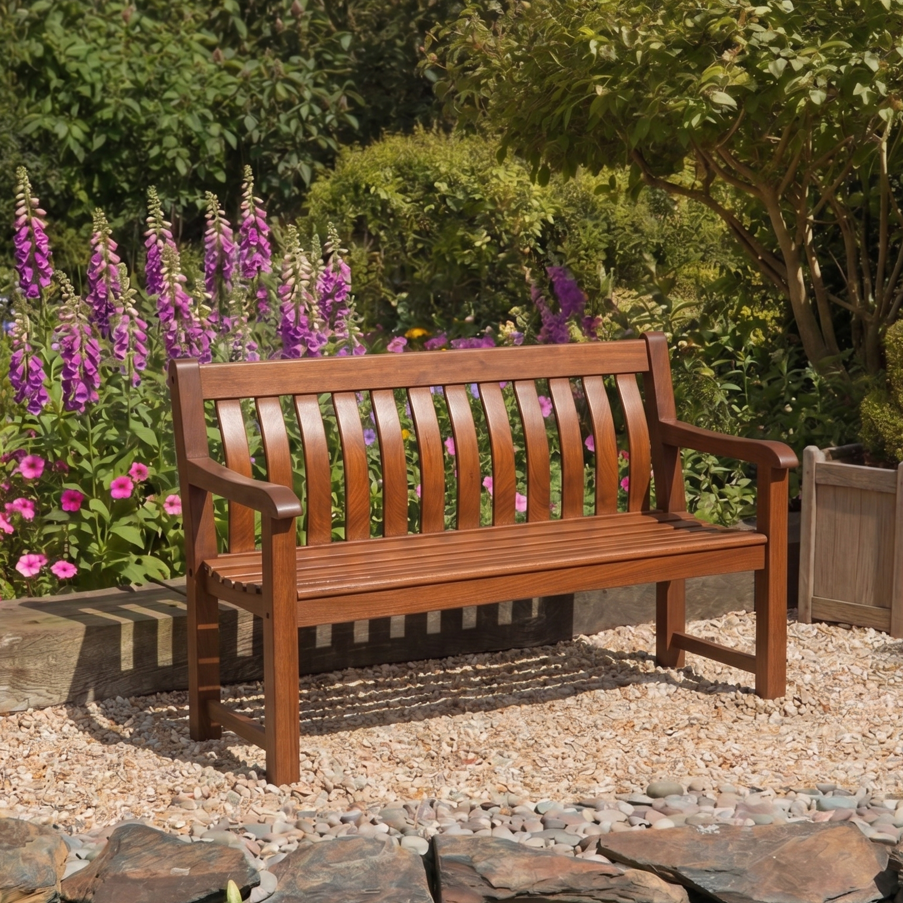 A wooden garden bench in mahogany with a slatted back and seat, placed on a gravel path in a landscaped garden with purple flowers and green foliage.