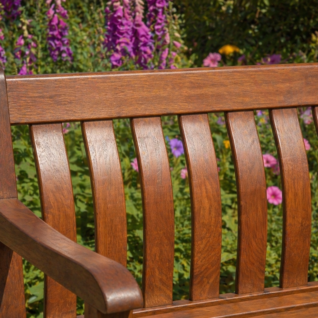 A close-up of a wooden garden bench in mahogany, set against a blurred background of green foliage and purple flowers.
