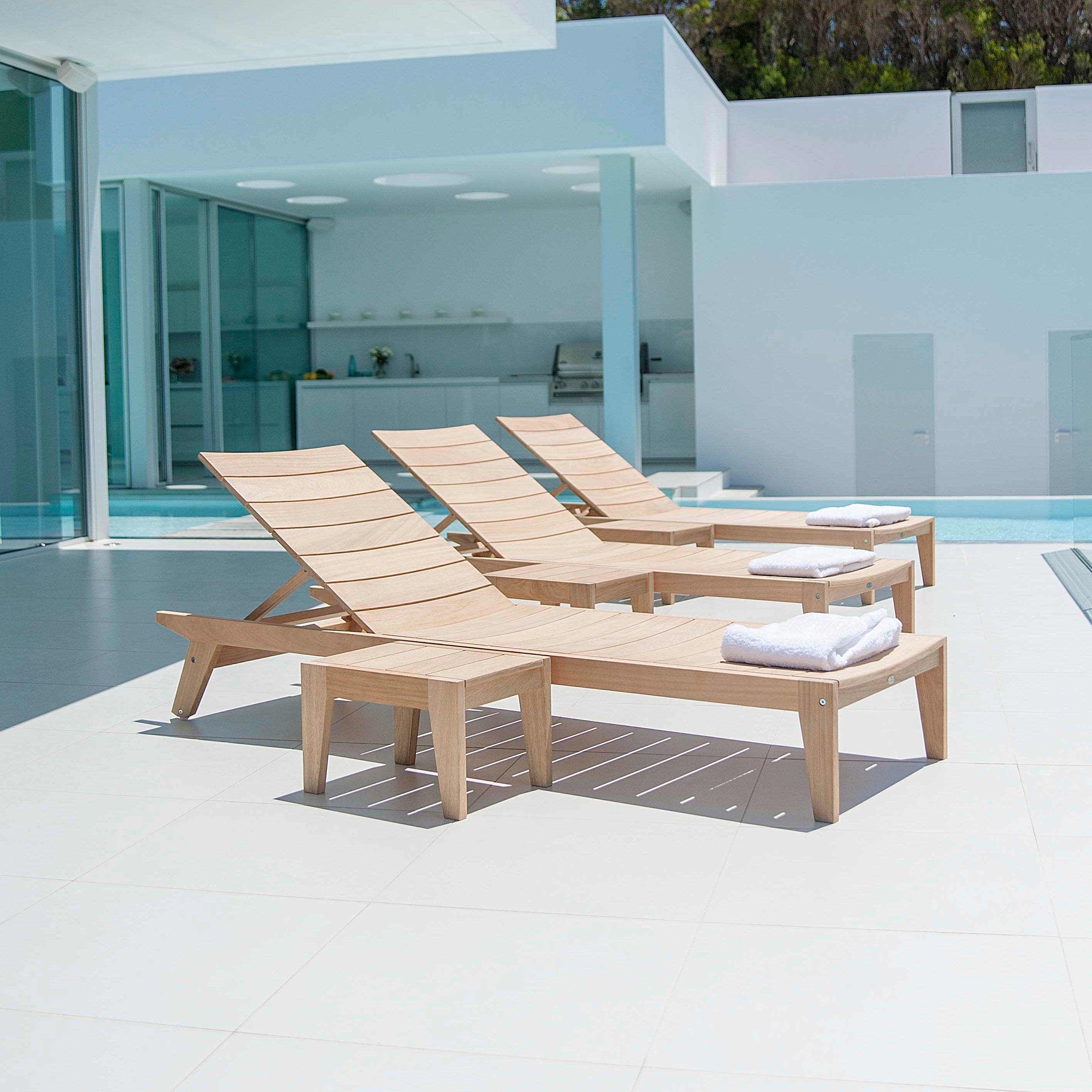 3 stylish roble wood sunbeds with side tables on a patio next to a pool