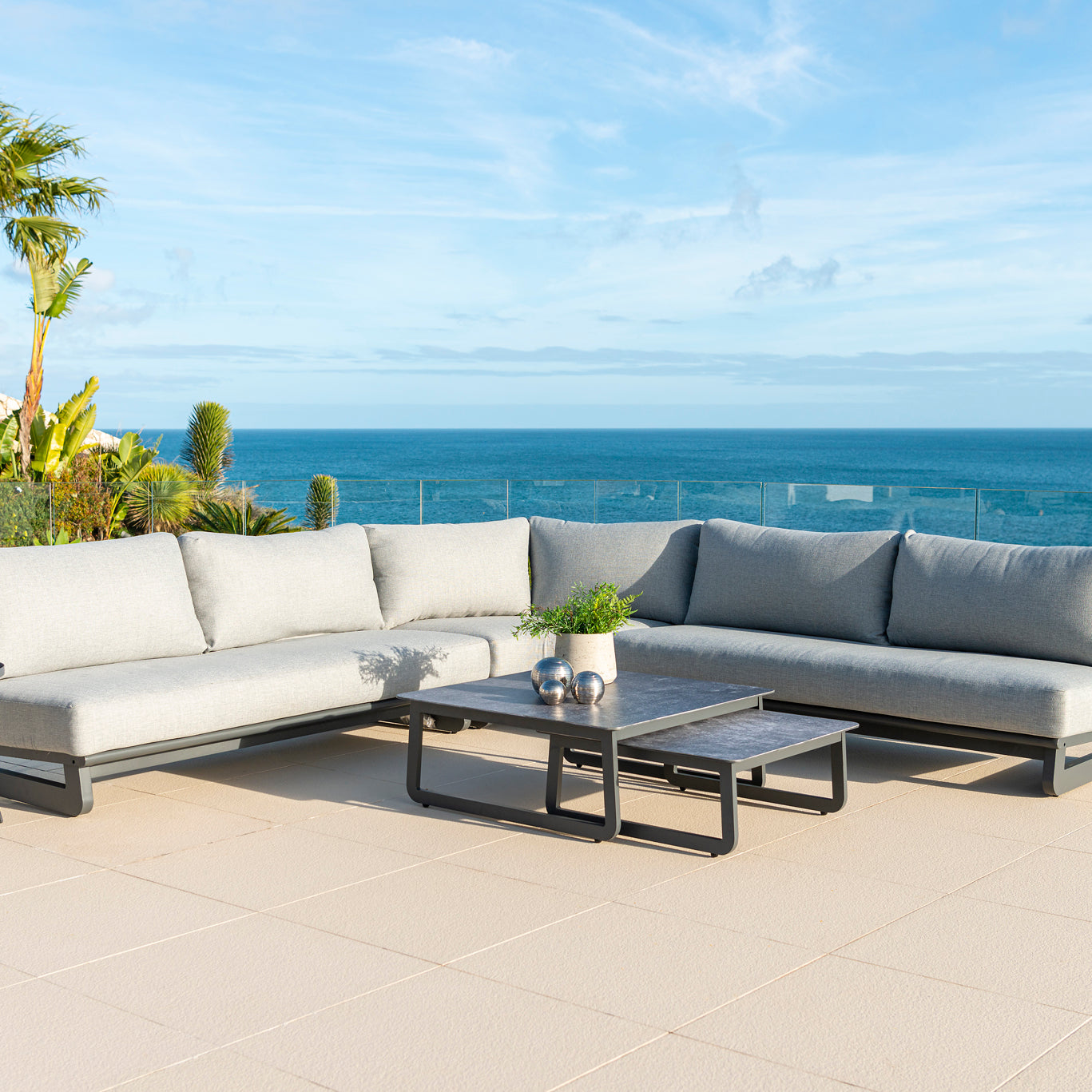 Rimini modular outdoor corner sofa set with light grey cushions, two coffee tables and bistro table on a patio with the sea in the background