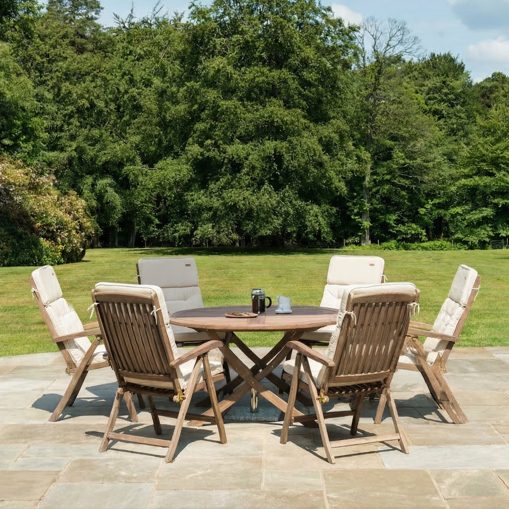 Outdoor patio set with wooden table and chairs on a stone patio with green grass and trees in the background.