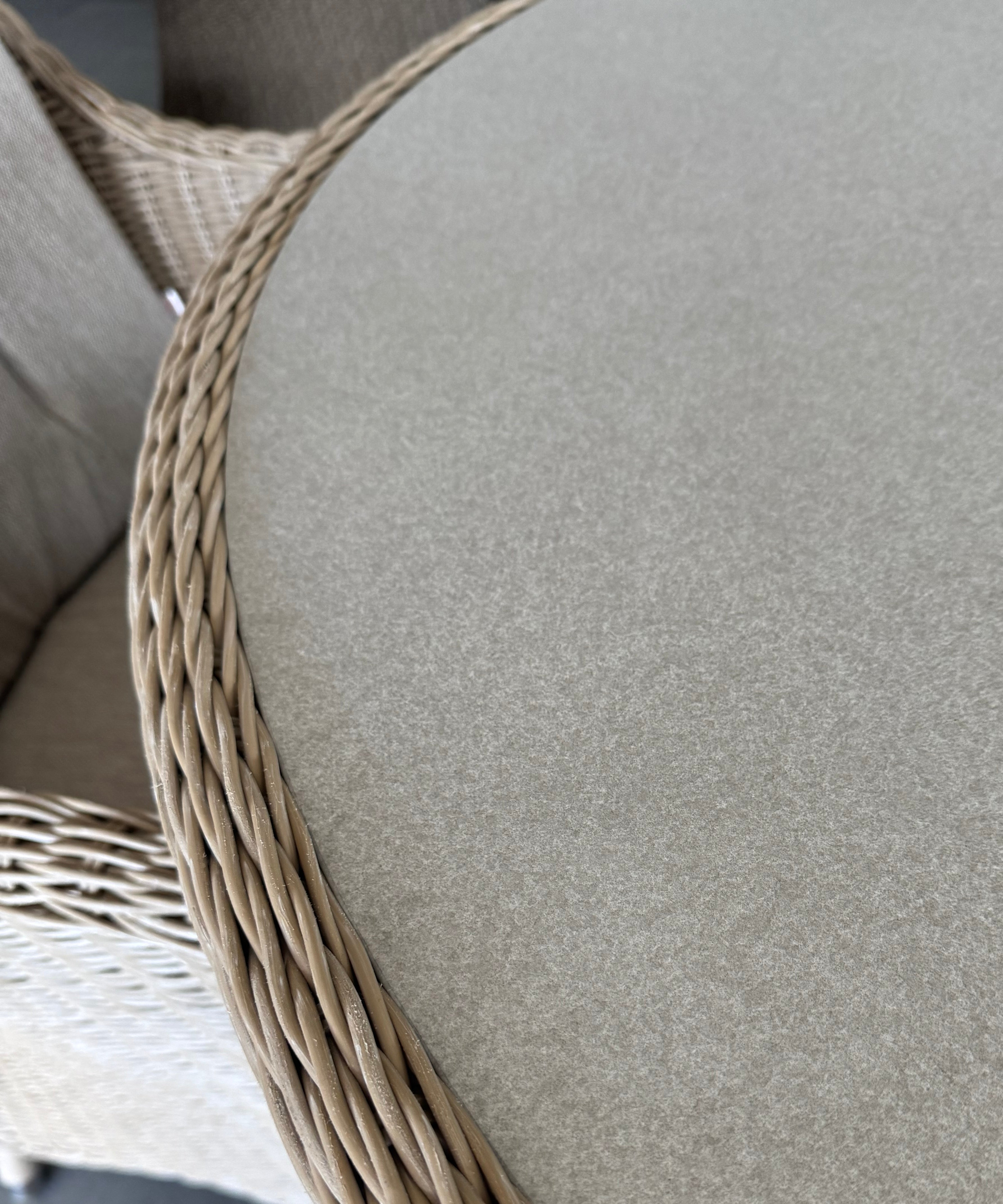 Close-up of a round outdoor dining table with a light beige woven wicker frame and a thick, textured beige fabric cushion.
