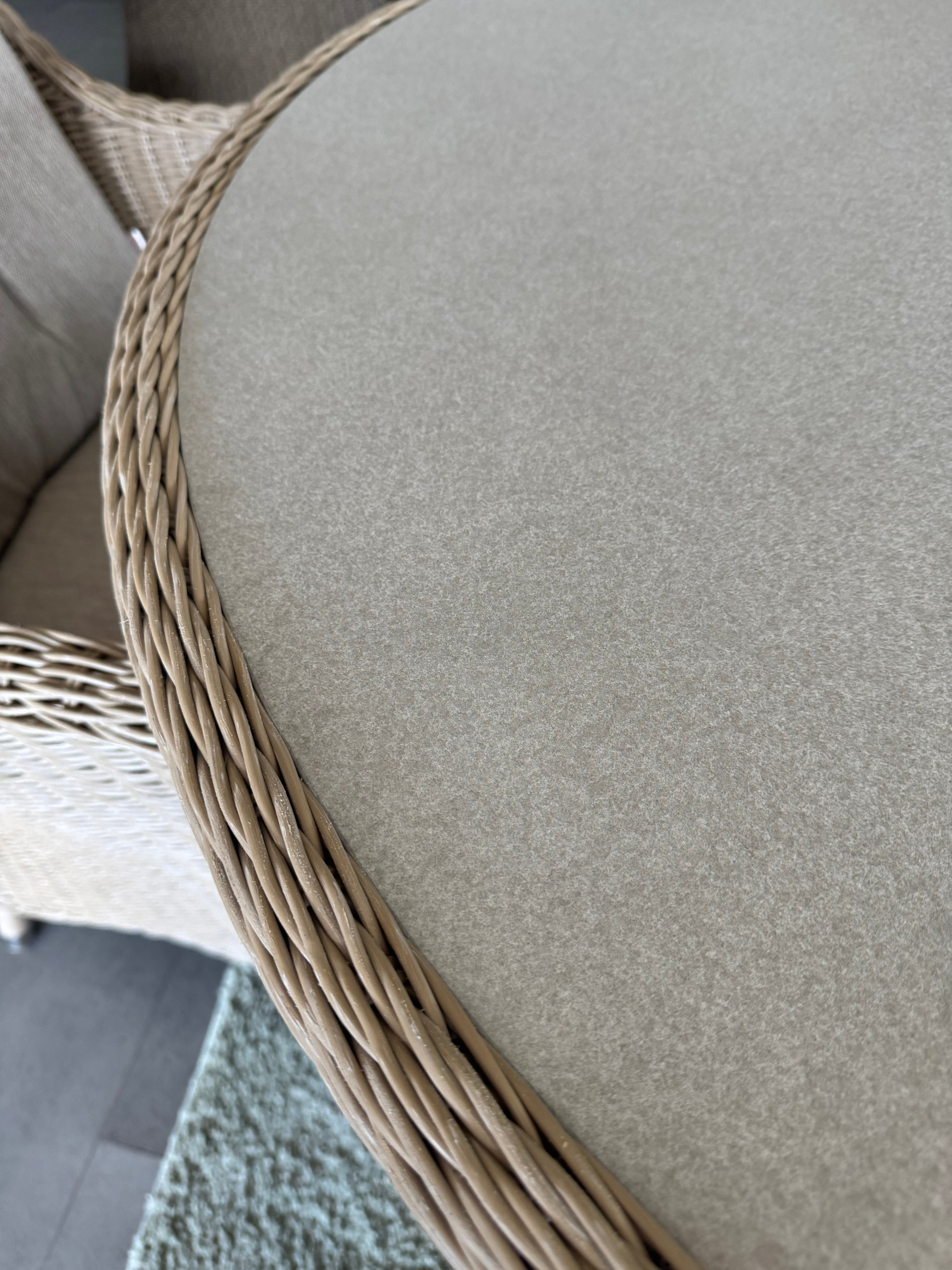 Close-up of a round outdoor dining table with a light beige woven wicker frame and a thick, textured beige fabric cushion.