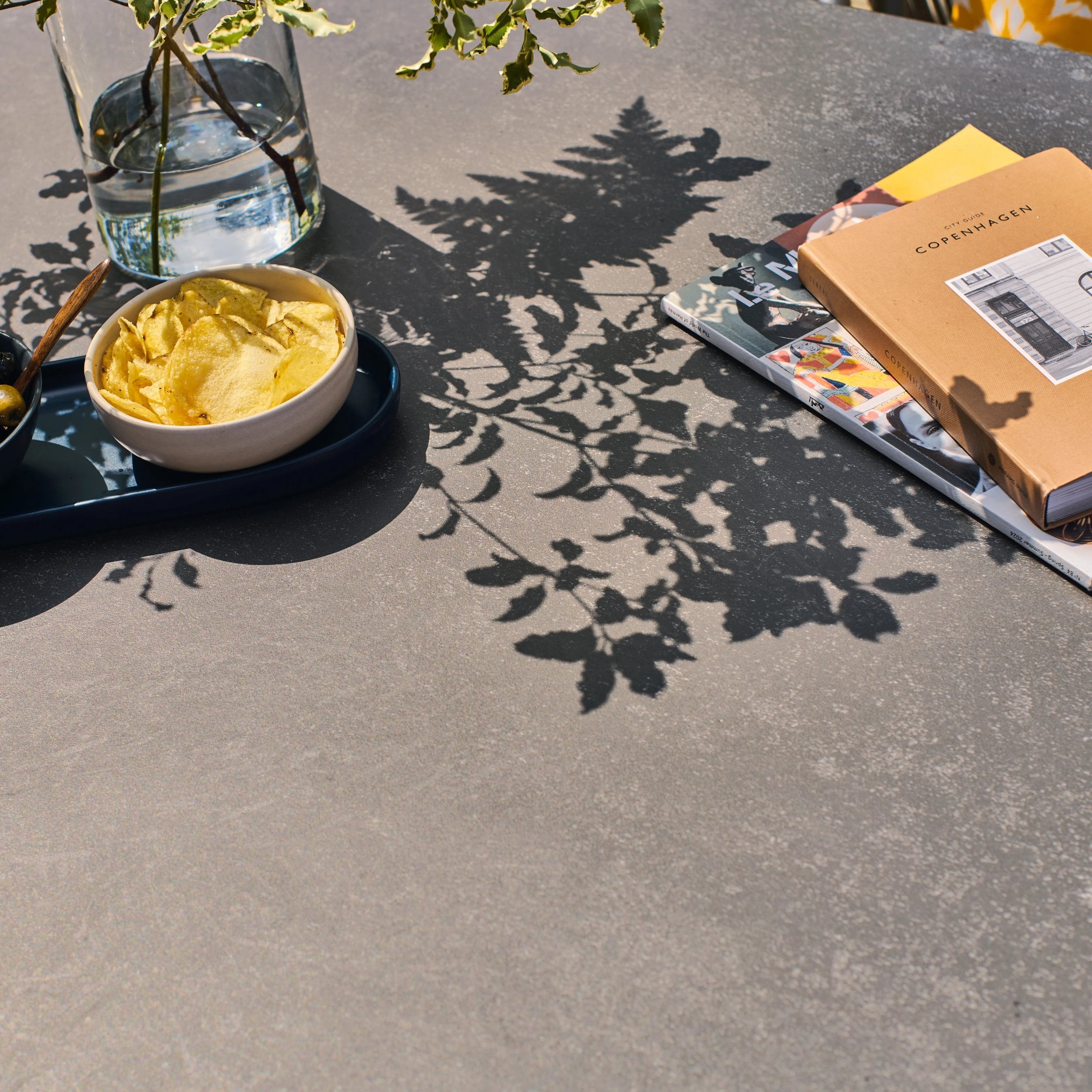 Bowl of chips on a tray with a notebook and plant shadows on a table top in grey colour