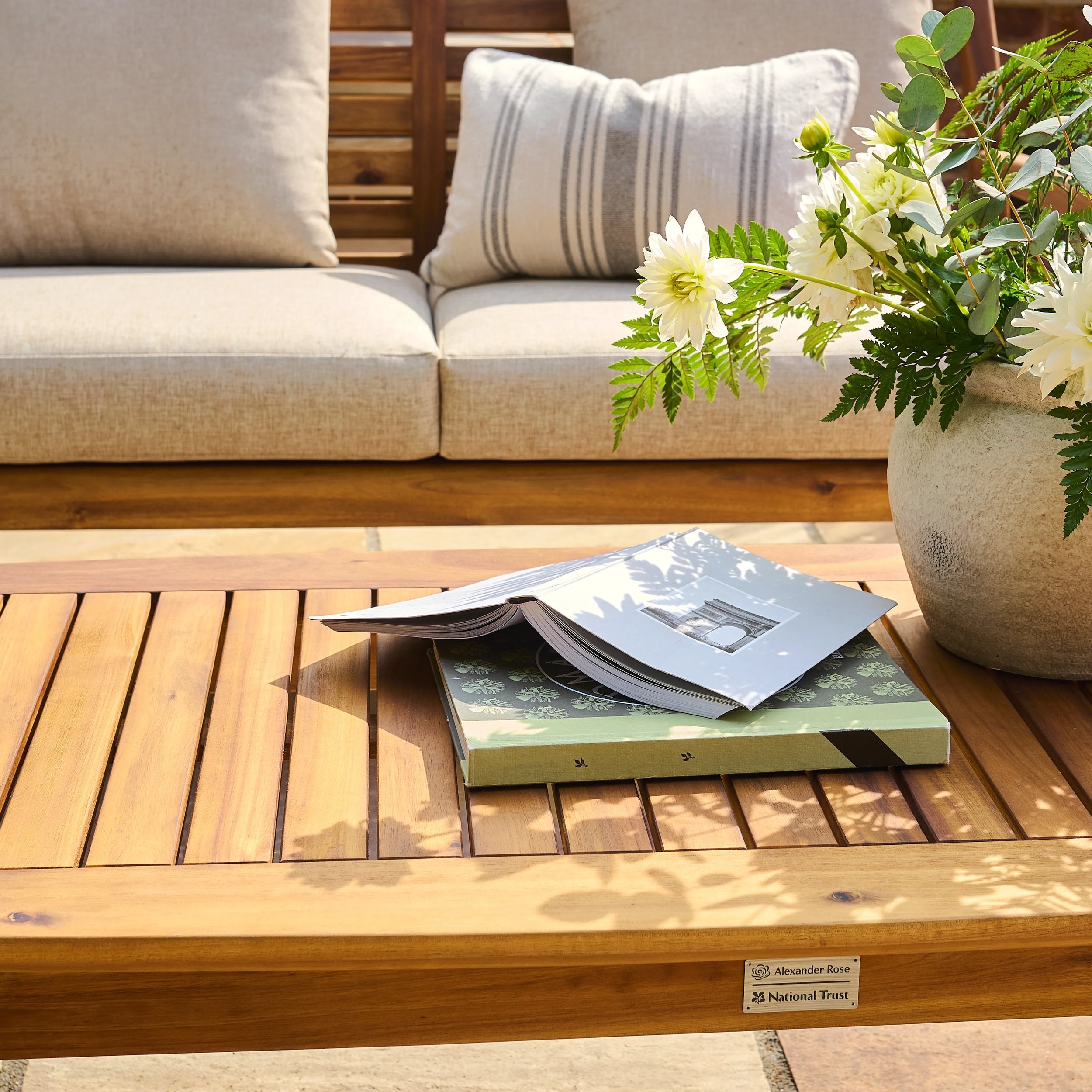 Wooden outdoor table with books and a vase of flowers, next to a beige sofa.