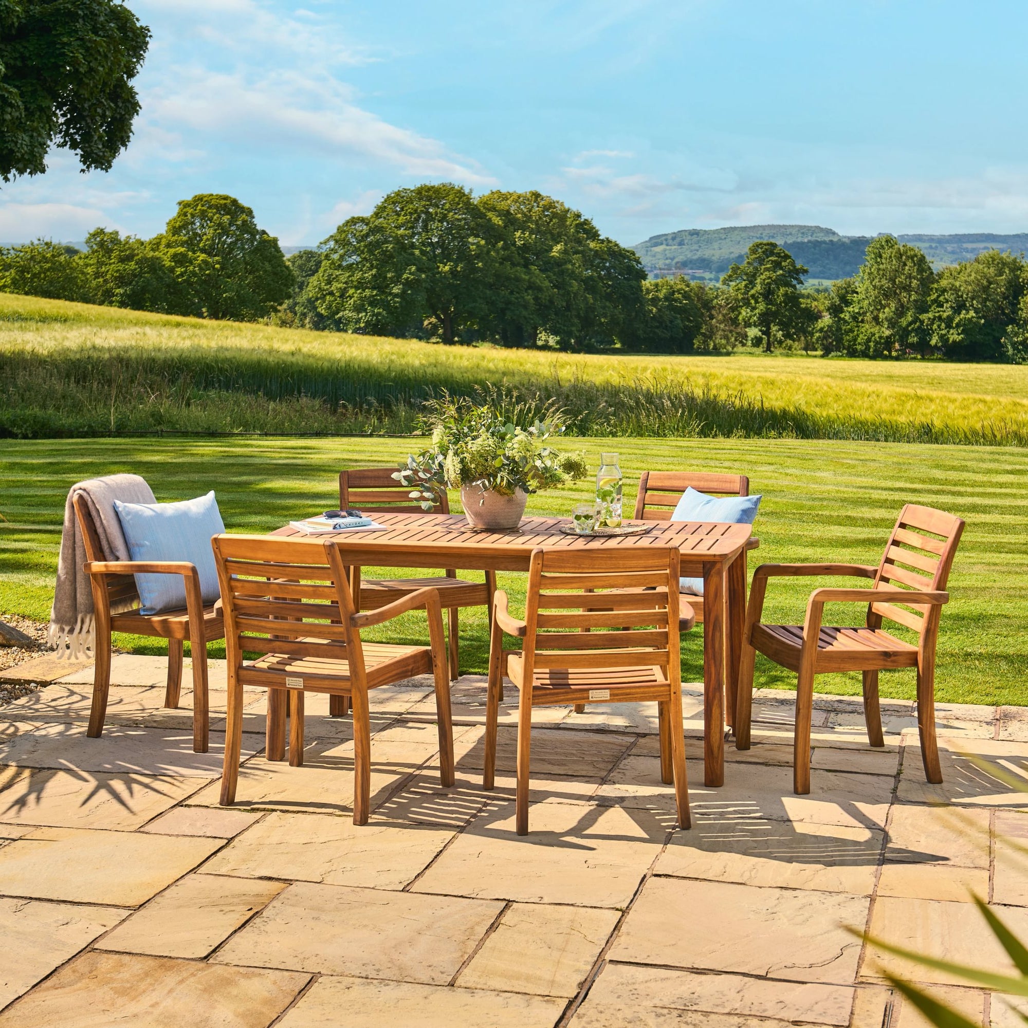 Outdoor dining set with wooden table and chairs on a patio, surrounded by greenery and mountains.