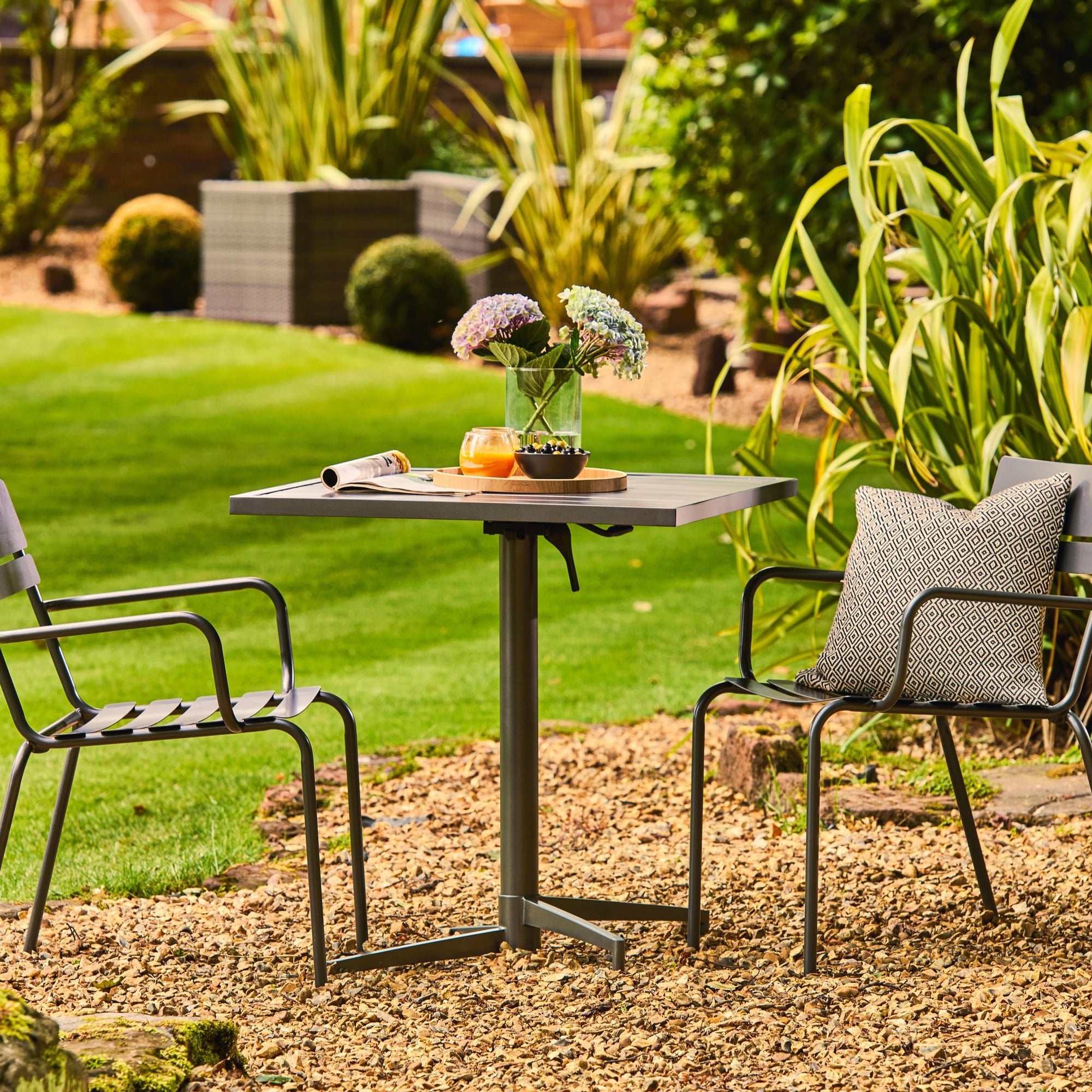Garden setting with a small table and two chairs on a gravel area, surrounded by greenery.
