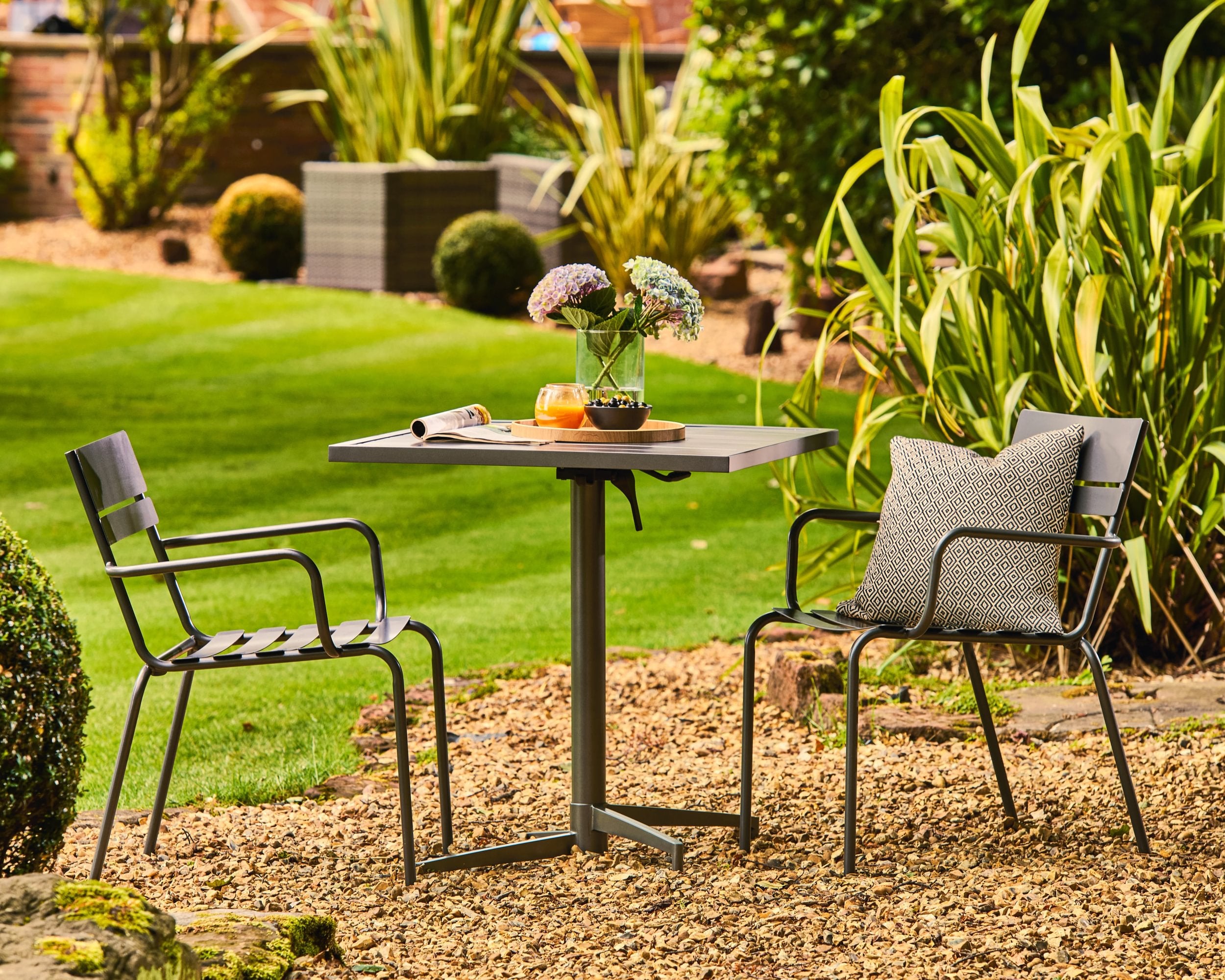 Garden setting with a small table and chairs on a gravel area, surrounded by greenery.