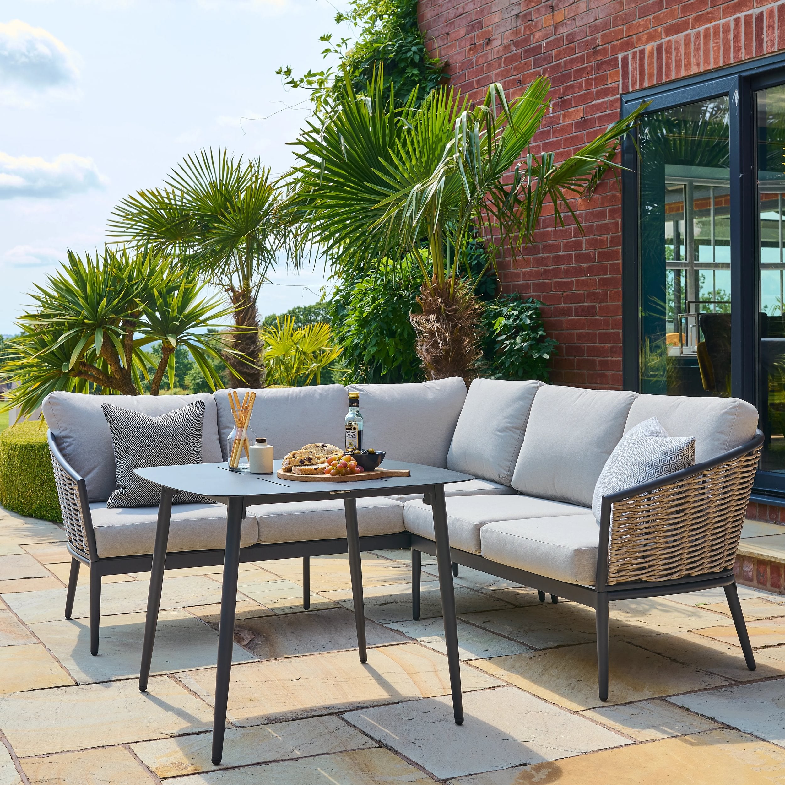 Outdoor patio set with gray cushions on a stone patio, surrounded by plants and a brick wall.