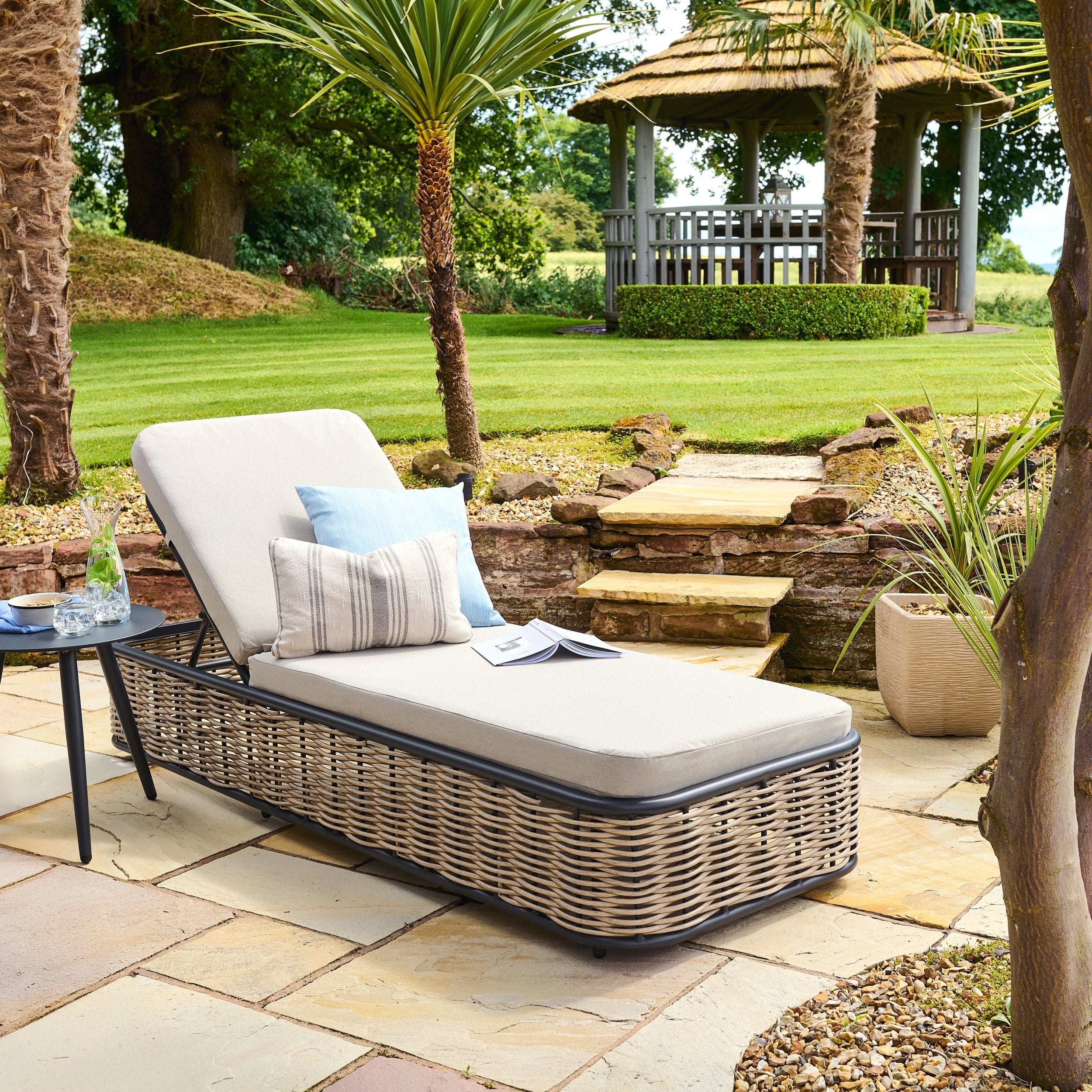 Rattan sun lounger with cushions on a stone patio in a garden setting
