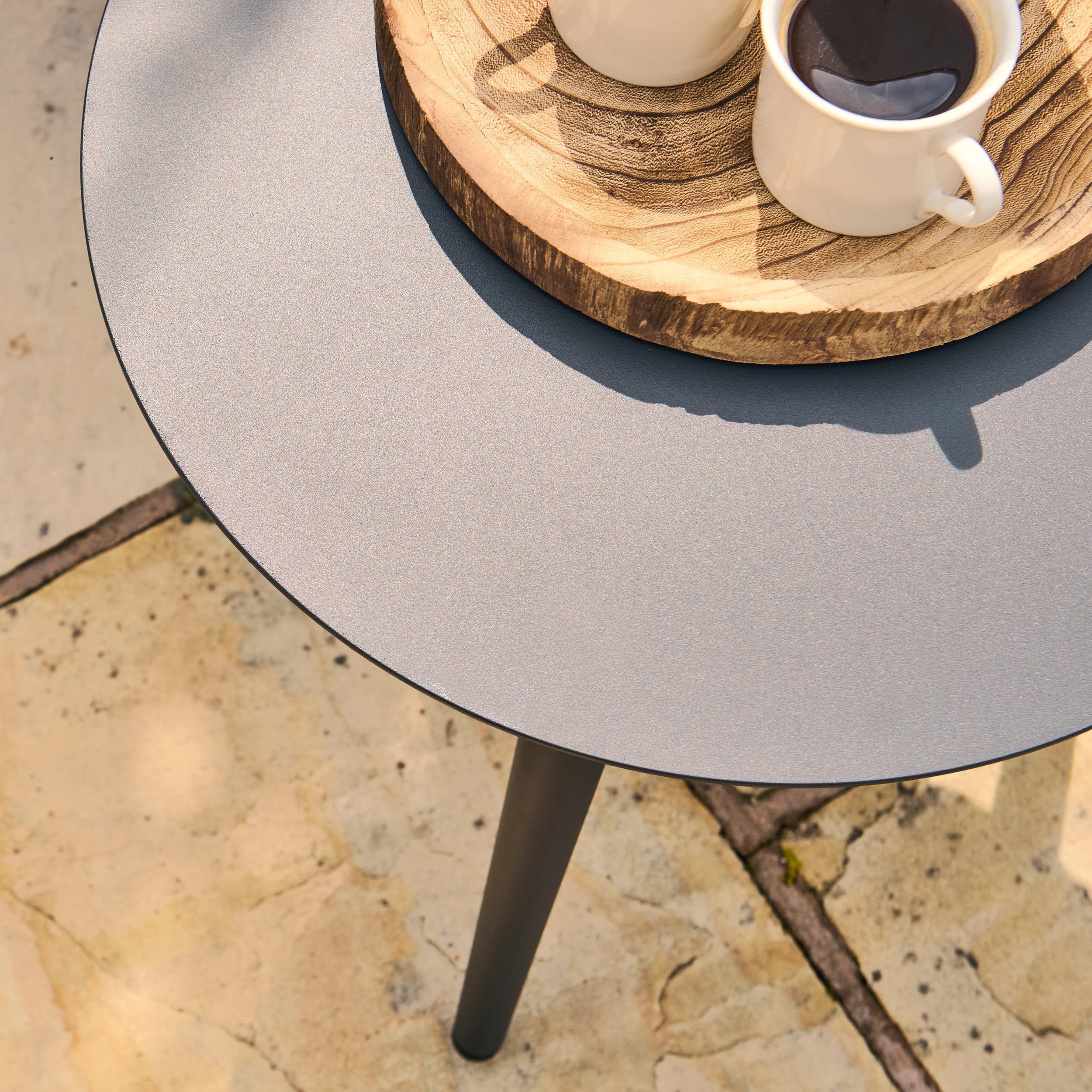 Round grey table with a wooden tray holding two white mugs on a stone patio.