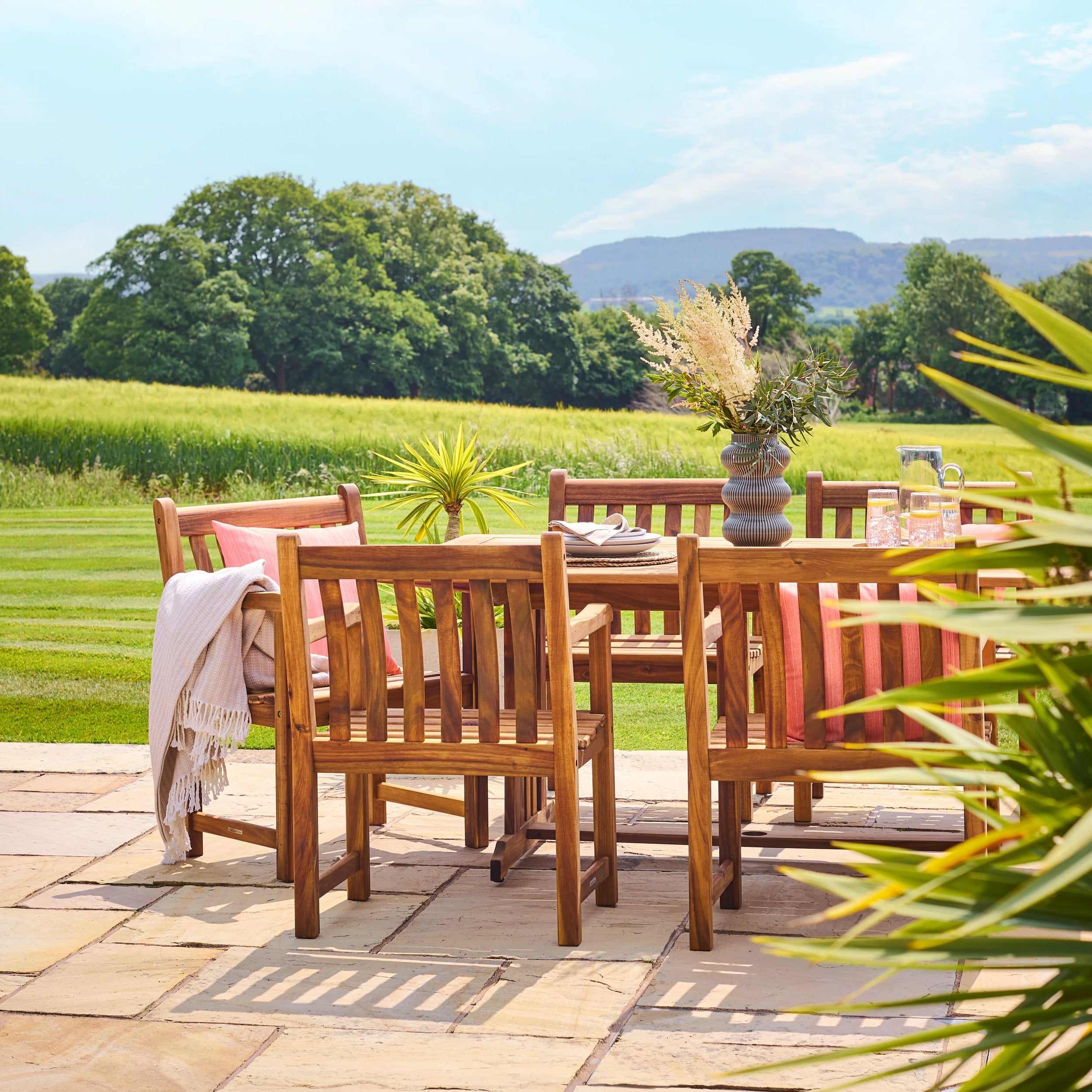 Wooden outdoor dining set with chairs and table on a patio, surrounded by greenery.