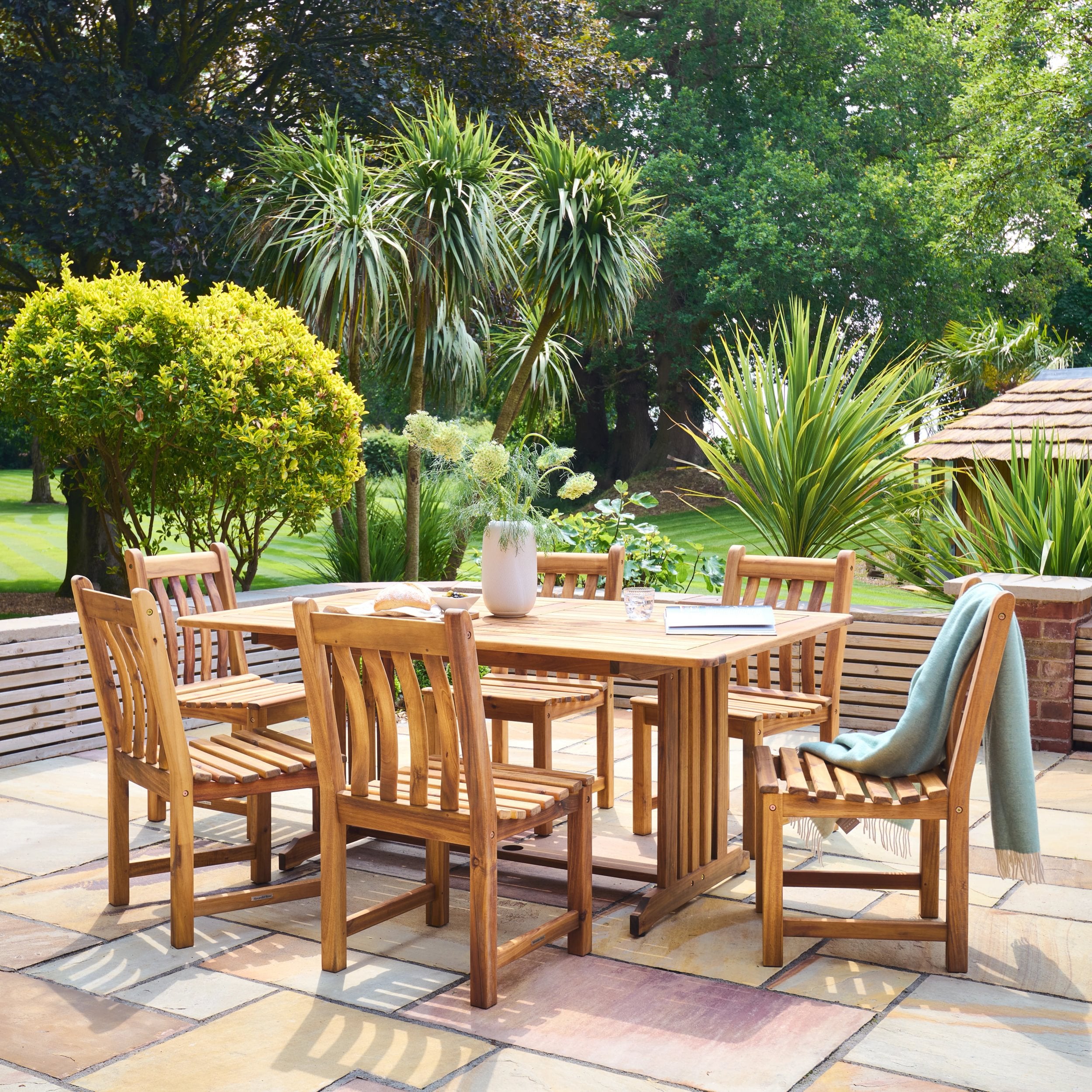 Wooden outdoor dining set with table and chairs on a patio, surrounded by greenery.