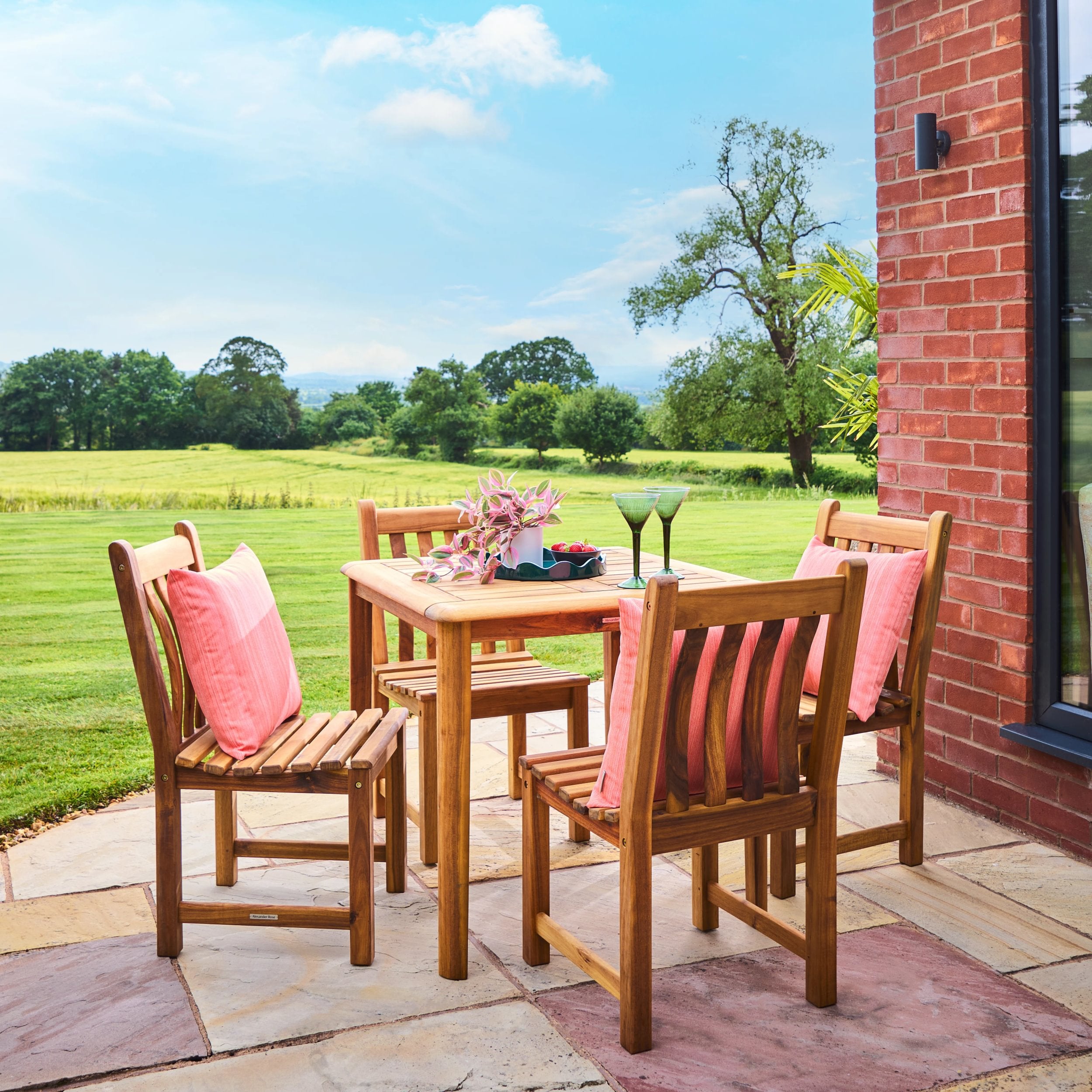 Wooden outdoor dining set with pink cushions on a patio with a view of green fields and trees.