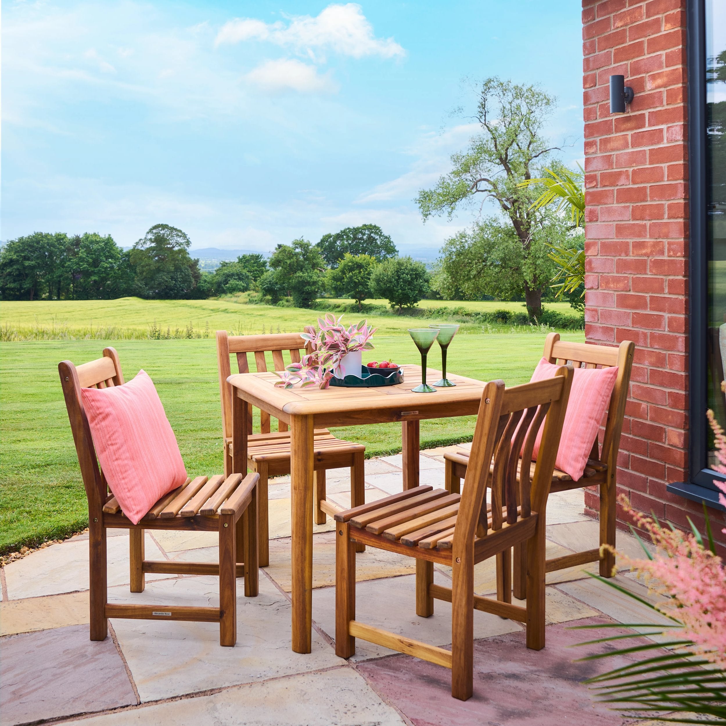 Wooden outdoor dining set with pink cushions on a patio with a view of green fields and trees.