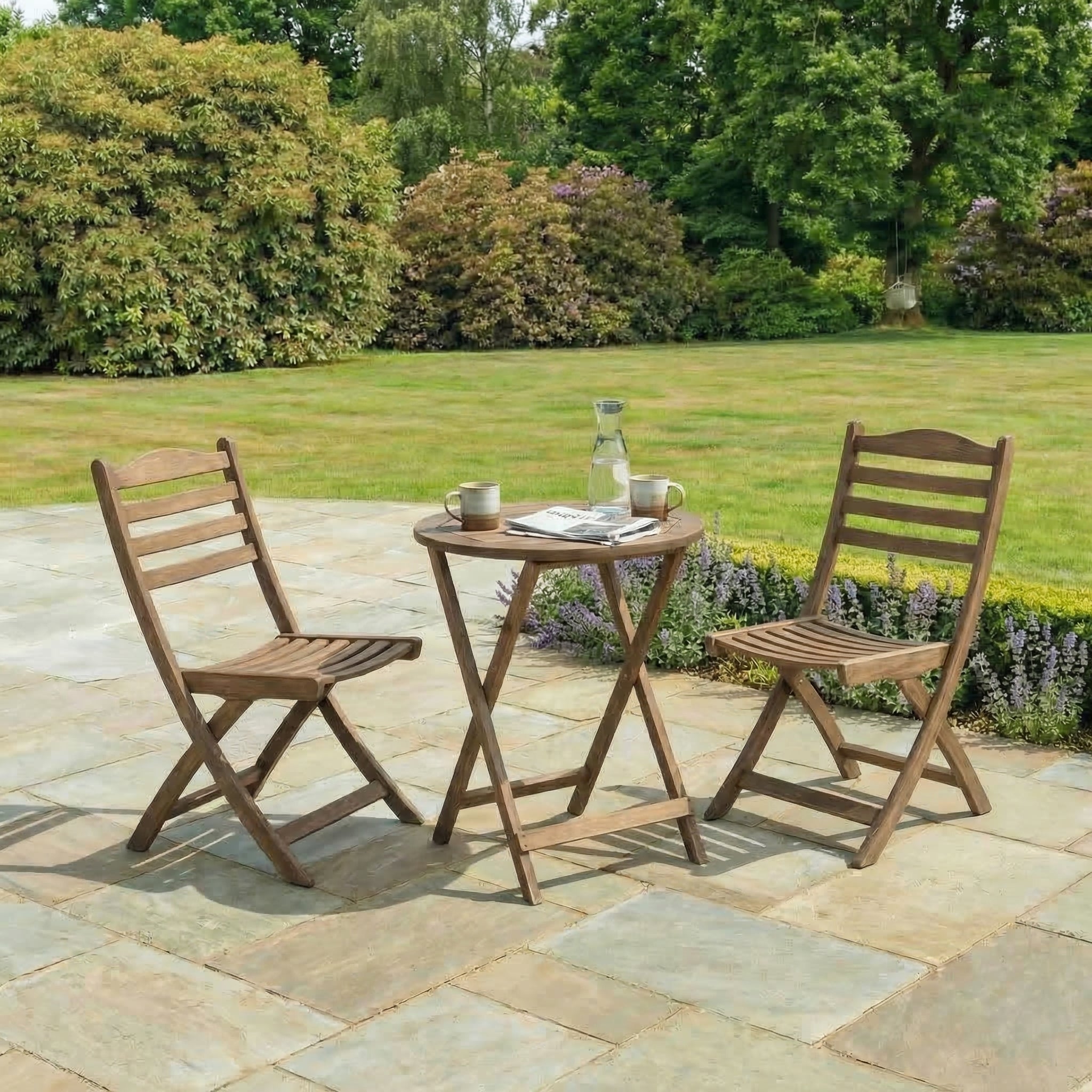 Wooden outdoor table with two chairs on a stone patio with greenery in the background