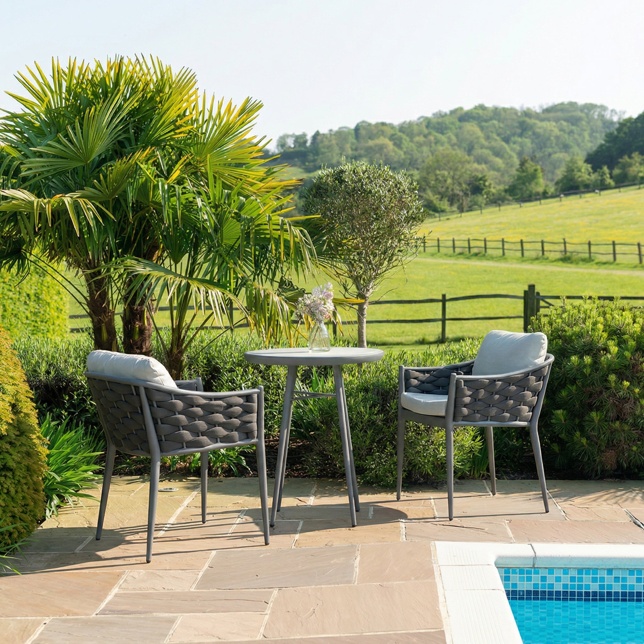Outdoor patio bistro set with table and chairs near a pool, surrounded by greenery.