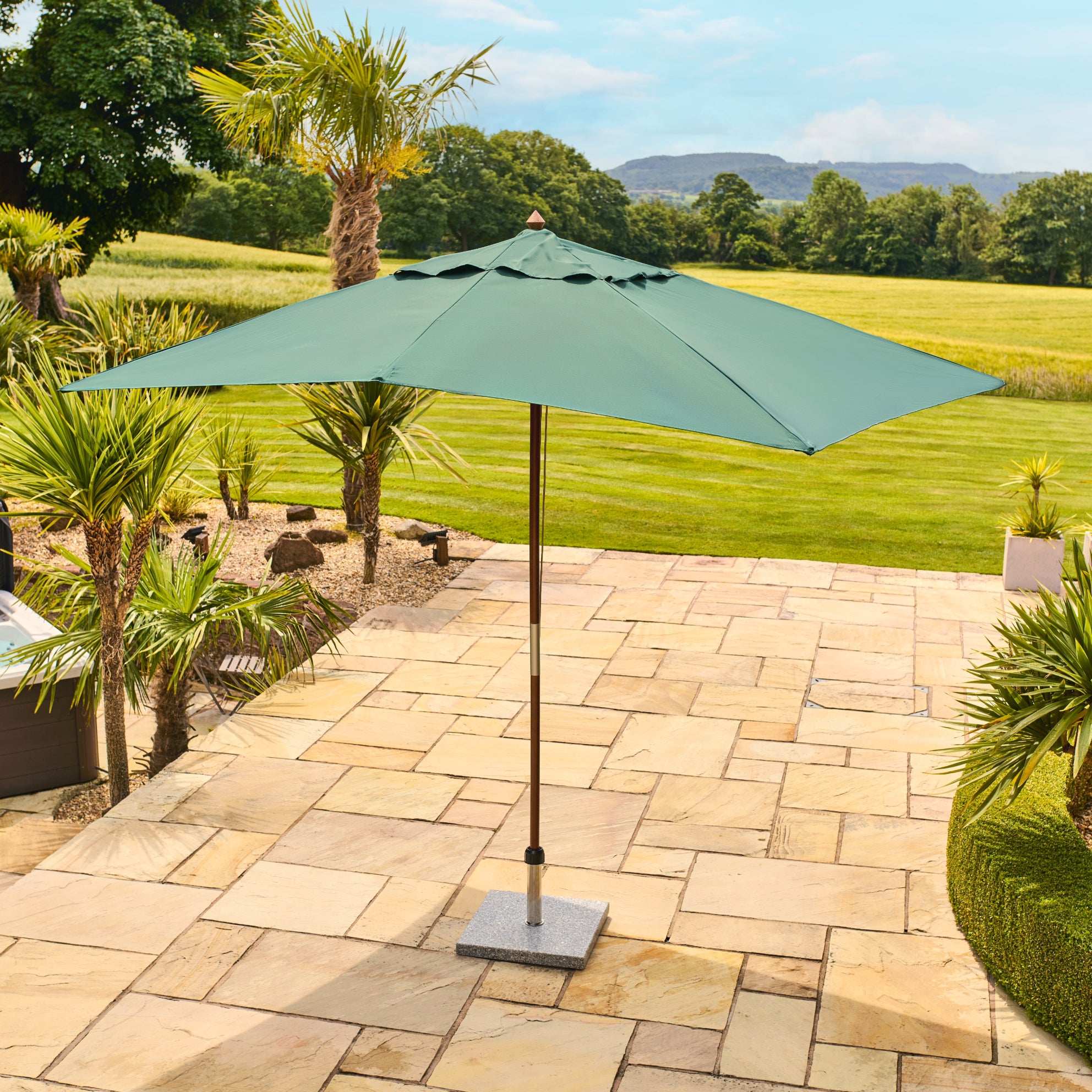 A green patio umbrella with a square canopy and central pole, standing on a stone patio in a landscaped backyard with grass, trees, and a hot tub nearby.